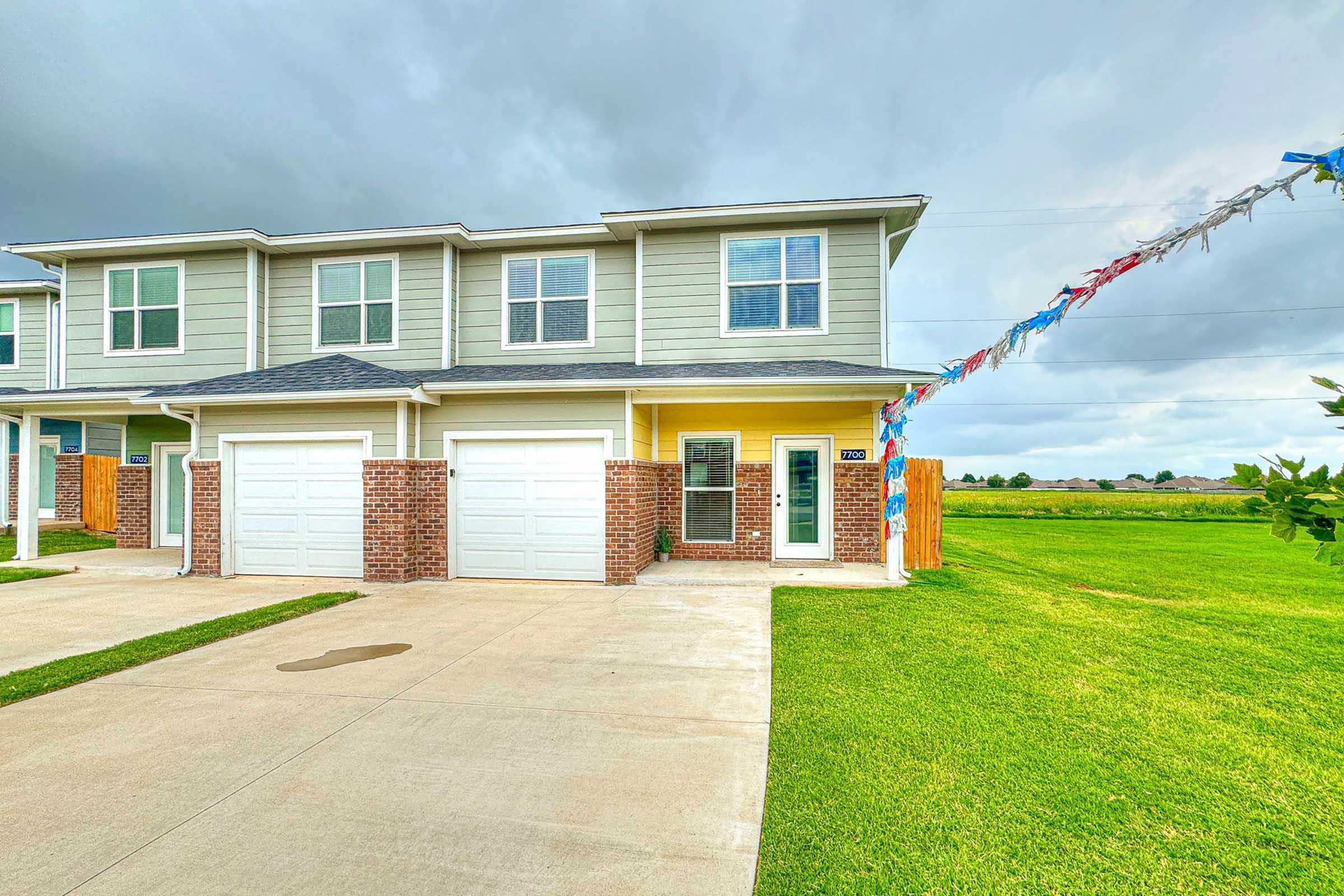 A two-story townhouse with light green siding and a brick exterior, featuring a garage and a front door with a window. The driveway is paved, with a green lawn extending to the left. Colorful decorative streamers are hanging from the eaves, and the sky is partially cloudy.