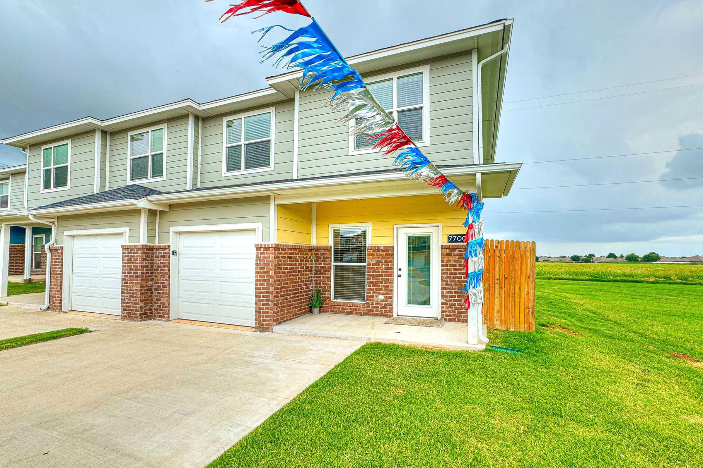 A two-story residential building with gray siding and brick accents. The building features two garage doors, a front entrance with a small porch, and is decorated with colorful banners. A green lawn surrounds the property, and a wooden fence provides privacy. The sky is partly cloudy.