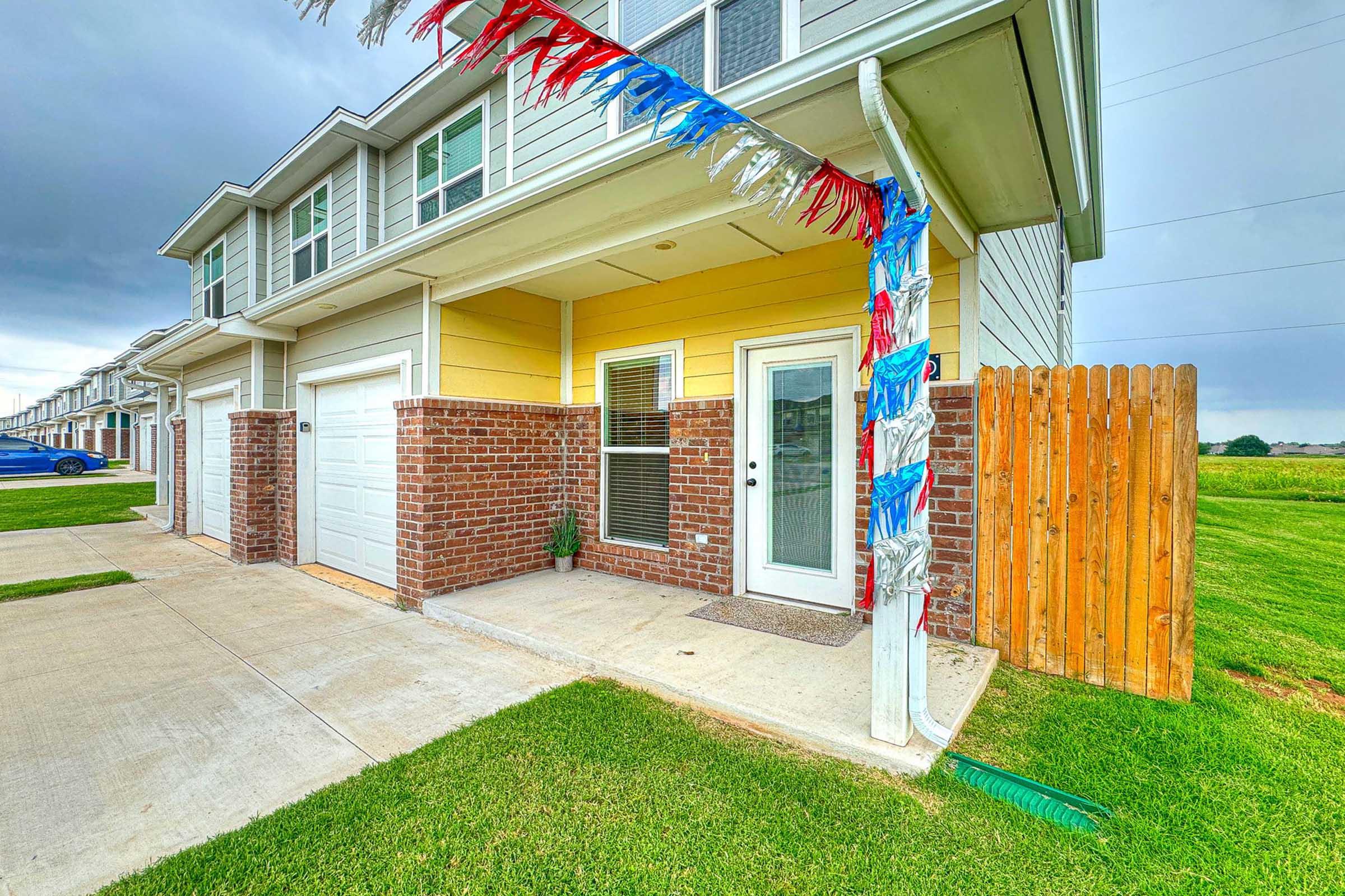 A residential building entrance featuring a yellow wall and a brick facade. There is a covered porch with a welcome mat and decorative red, white, and blue streamers hanging from the roof. The area is neatly landscaped with green grass and a wooden fence in the background.