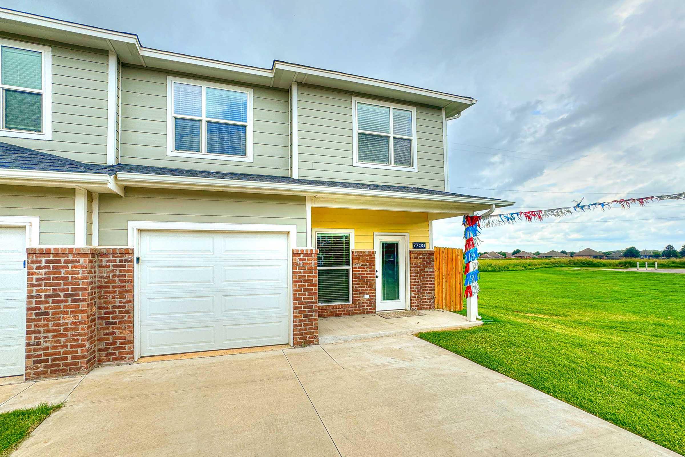 A modern two-story house with a light green exterior, white trim, and a garage door. The front yard features a neatly paved driveway leading to the entrance. Colorful decorations are hung from the porch, and a spacious lawn extends into the distance under a cloudy sky.