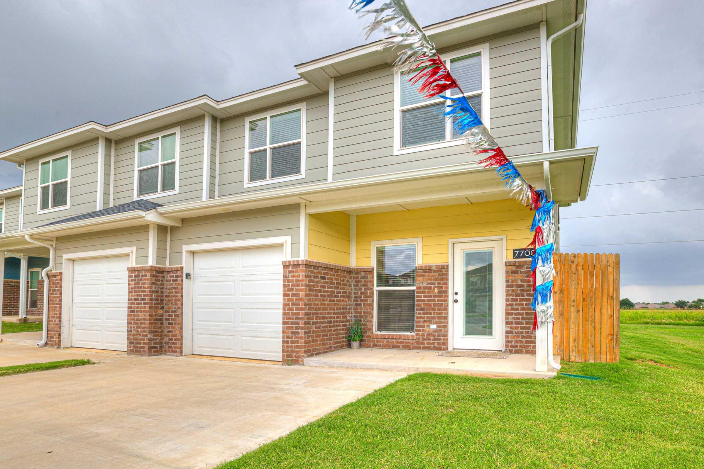 A two-story residential building featuring a yellow and gray exterior, with brick accents. The entrance has a front door and windows, and there's a small lawn in front. Decorative red, white, and blue streamers hang overhead, adding a festive touch to the scene.