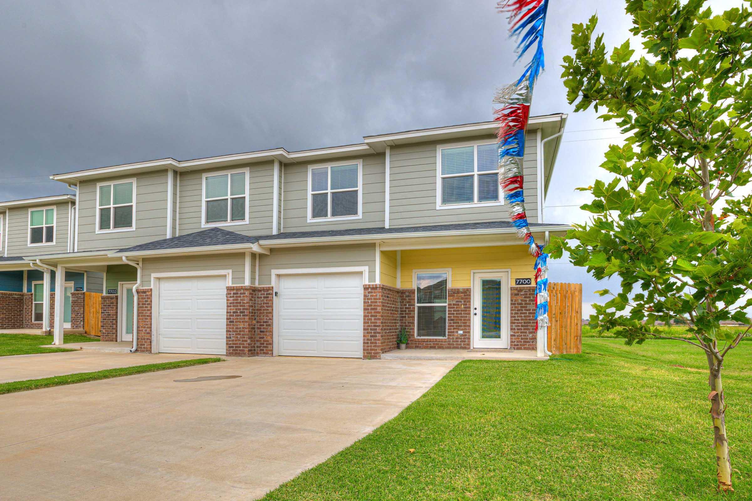 A two-story residential building with a modern design, featuring a grey and yellow exterior, and two garage doors. The front yard is well-maintained with grass, and decorative streamers are hanging from a nearby tree. There are dark clouds in the sky, indicating an overcast day.