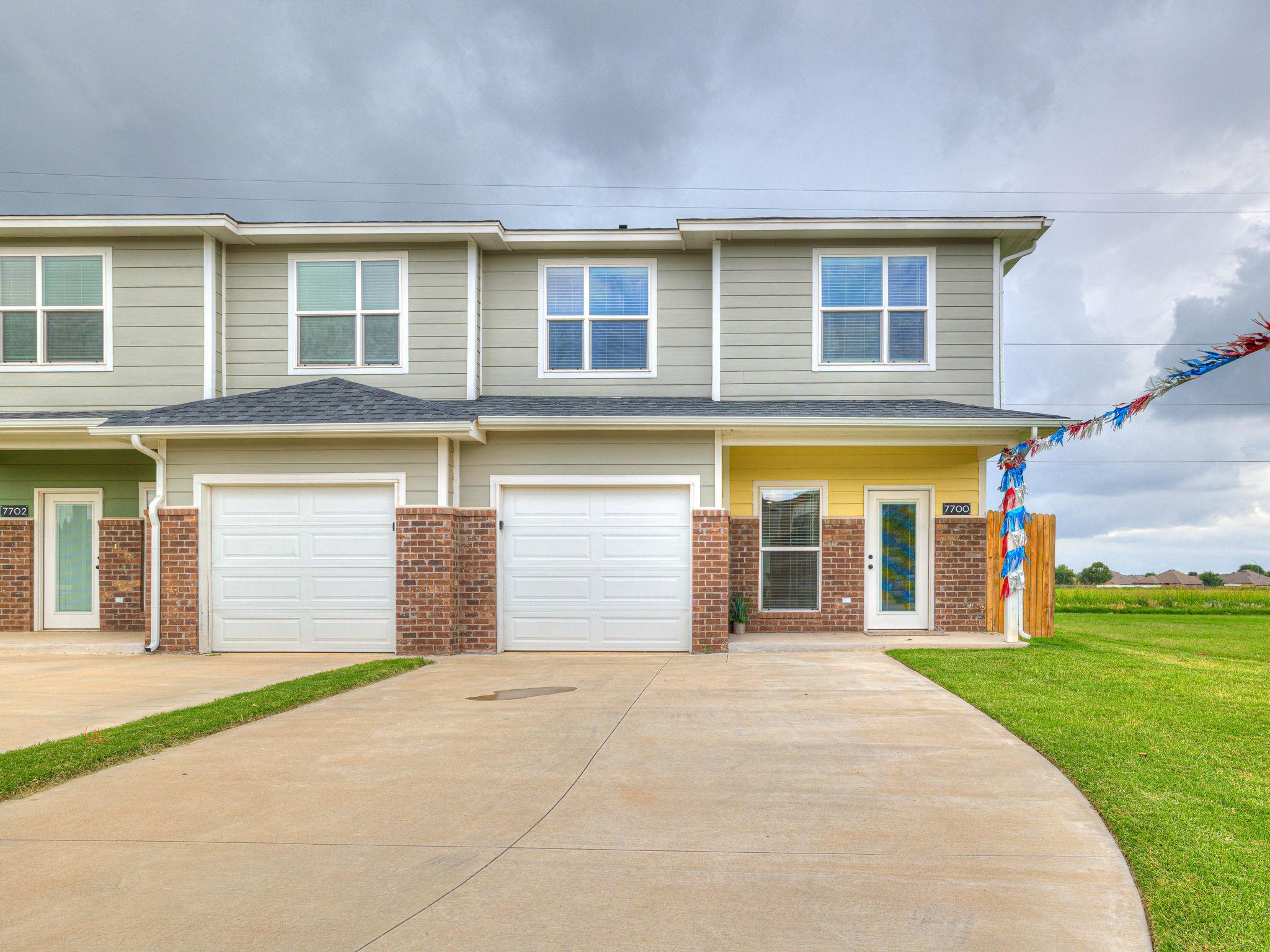 A two-story townhouse with a light gray exterior and a front porch. The building features two garage doors, large windows, and decorative elements like a colorful banner. A well-maintained lawn and open field can be seen in the background under a cloudy sky.