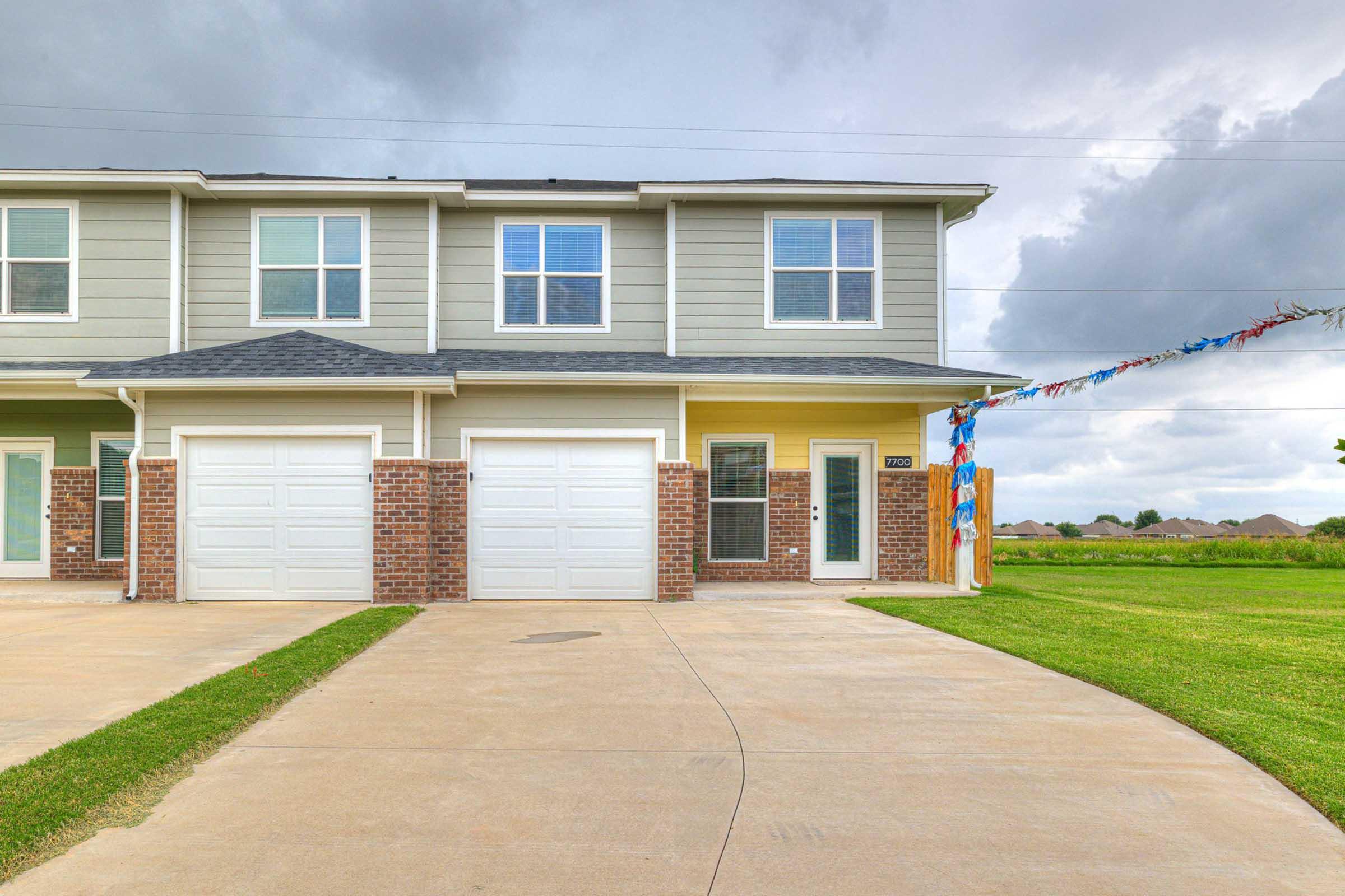 A residential townhouse featuring a two-car garage, with a light gray exterior and dark brick accents. The driveway is concrete, and there are large windows on the upper floor. The surrounding area is open with a grassy field visible in the background under a cloudy sky.