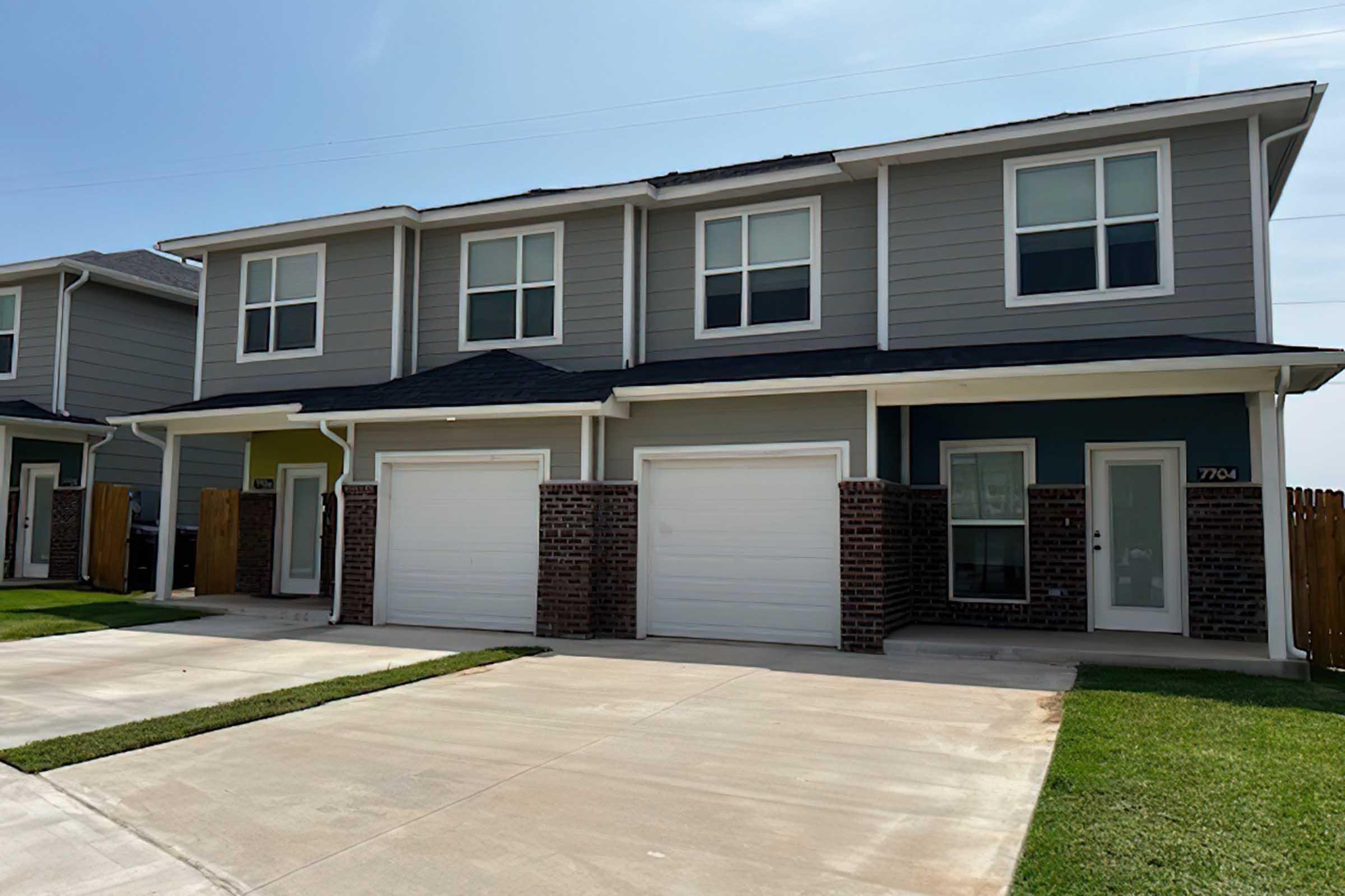 Two attached townhouses with gray siding and a brick base, featuring white garage doors. The facade includes multiple windows and a small porch area. The driveways are paved with concrete, and there is a grassy area in front of the buildings. Clear blue sky is visible above.
