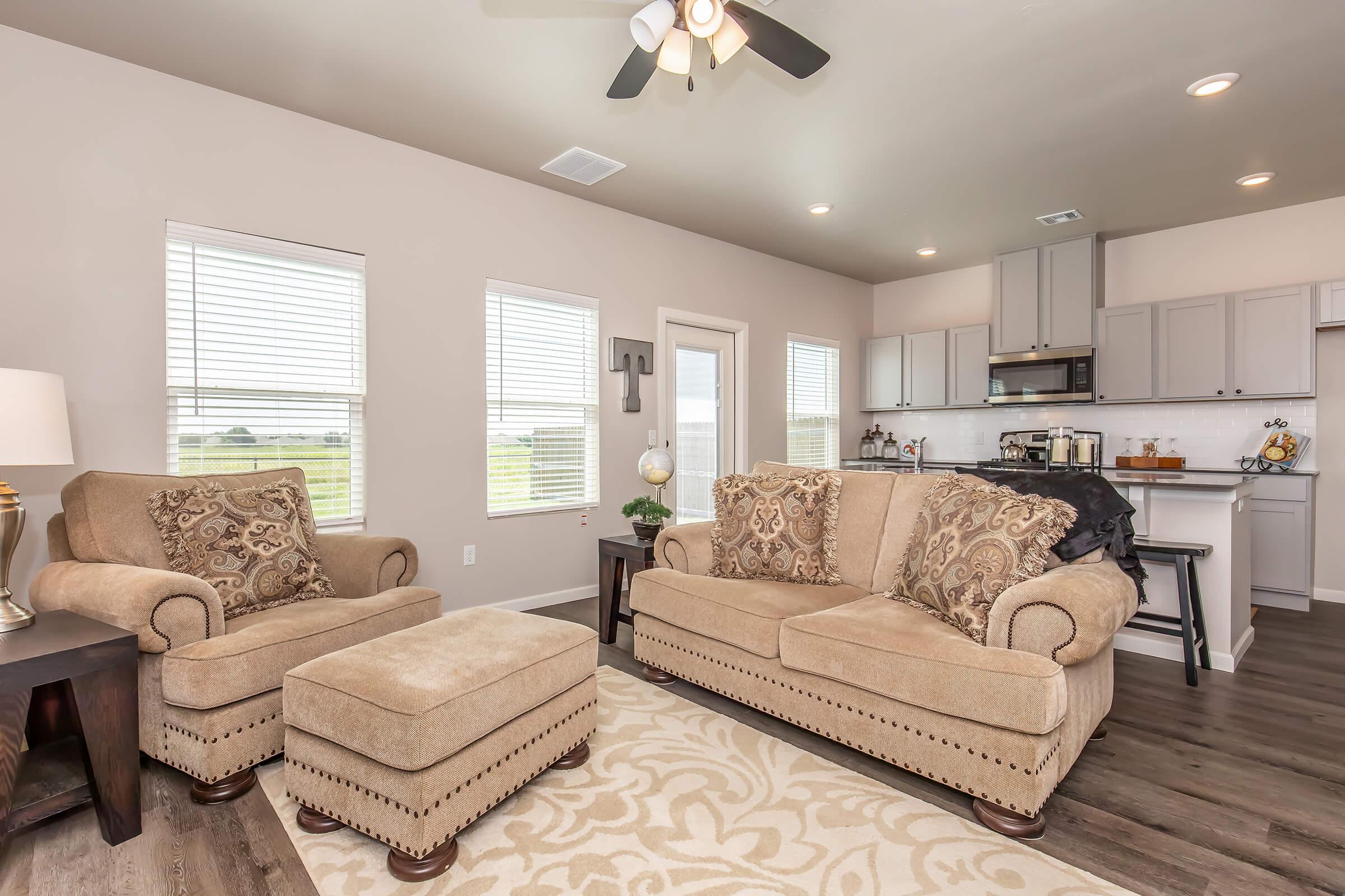 Cozy living room featuring two beige patterned armchairs and a matching ottoman, a small wooden side table, and a light fixture. The space has light-colored walls, large windows letting in natural light, and a view of the kitchen area with modern appliances and cabinetry in the background.