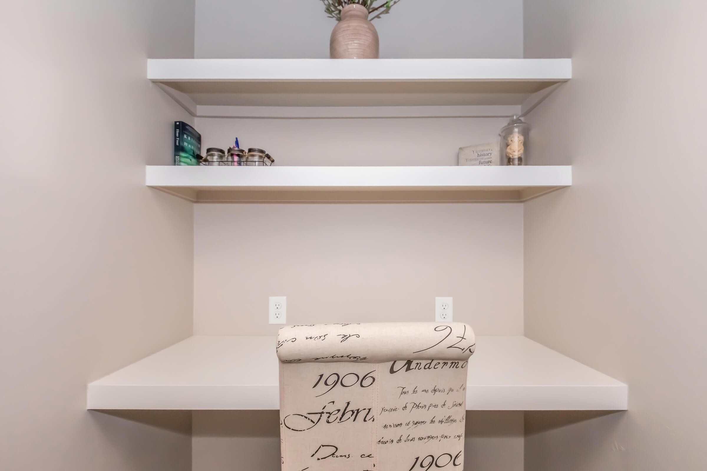 A minimalist workspace features a simple white desk with a patterned chair. Above, two floating shelves hold decorative items, including a vase with flowers and various jars. The wall is a neutral color, creating a calm and organized environment.