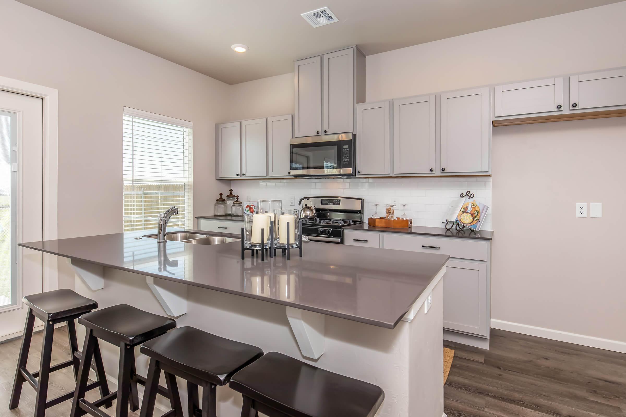 Modern kitchen featuring gray cabinetry, a stainless steel microwave, and gas stove. The island includes a gray countertop and four black stools. Natural light filters in through a window, highlighting a minimalist decor with candles and decorative items on the counter.