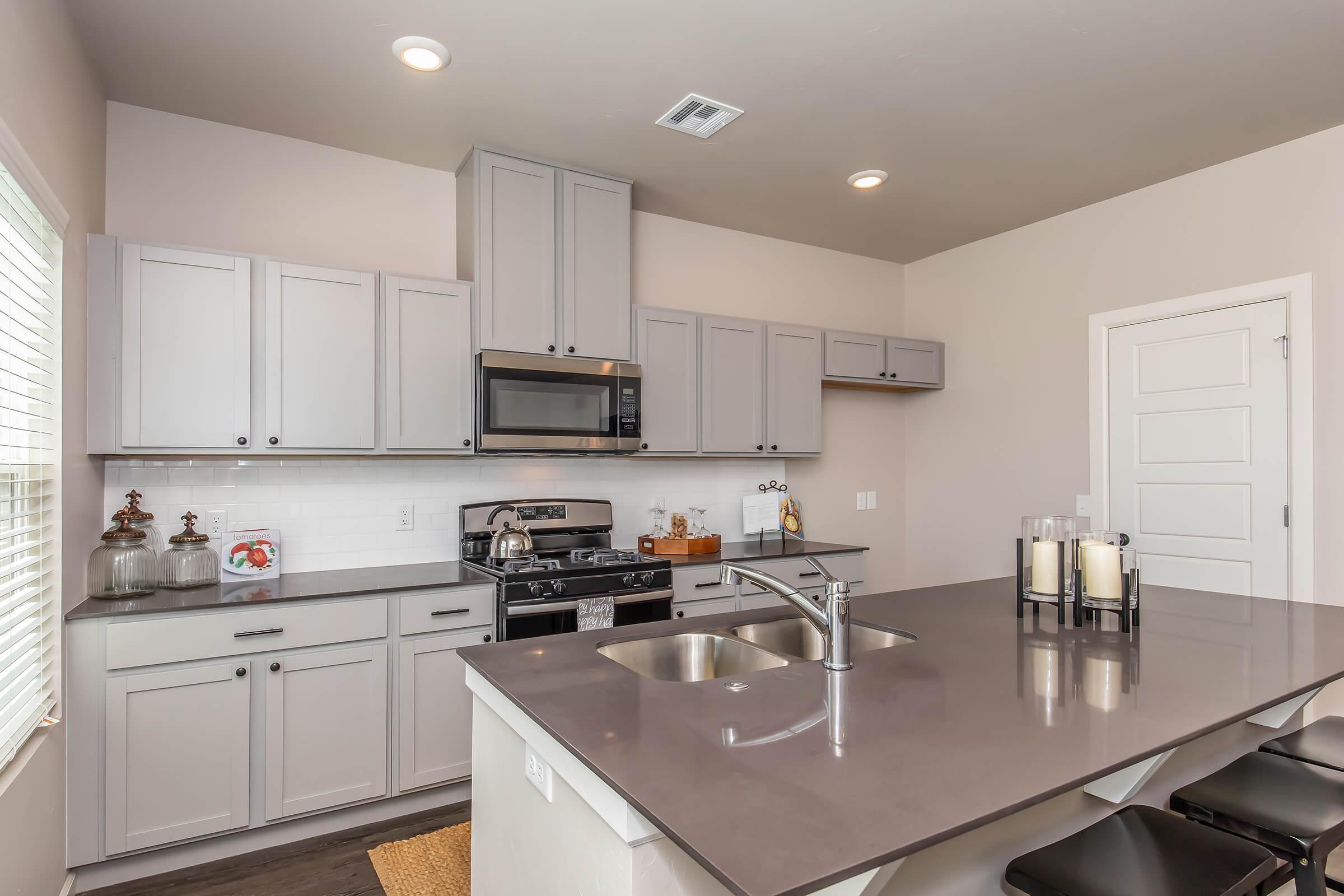 Modern kitchen featuring gray cabinets, stainless steel appliances, and a large island with dark countertops. The space is well-lit with natural light coming through a window, and there are bar stools at the island. Decorative items are placed on the countertops, adding a welcoming touch.
