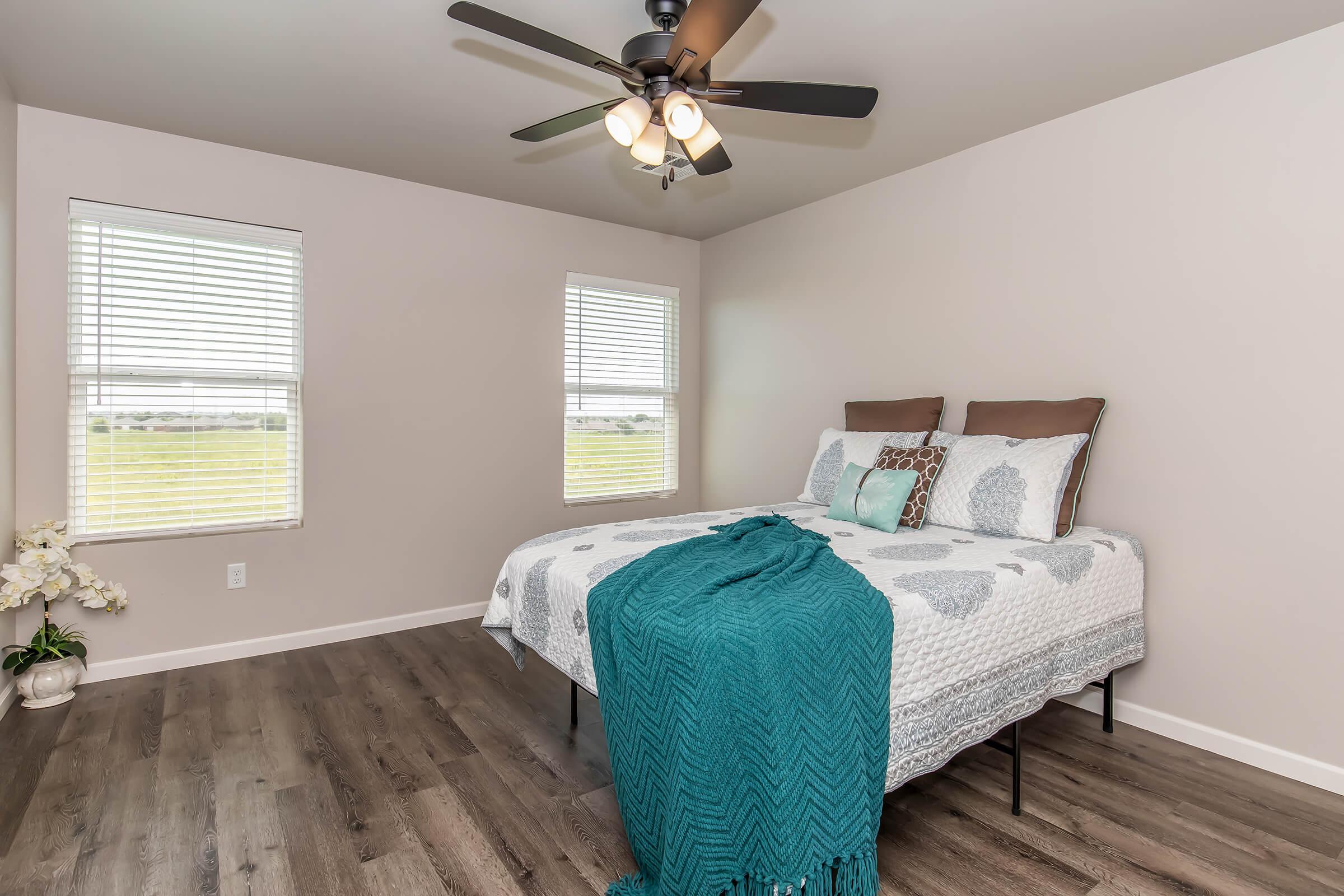 A cozy bedroom featuring a bed with a decorative quilt and pillows, two windows with white blinds letting in natural light, and a ceiling fan. There is a teal throw blanket draped over the bed and a small plant in the corner, highlighting a modern and inviting decor style.