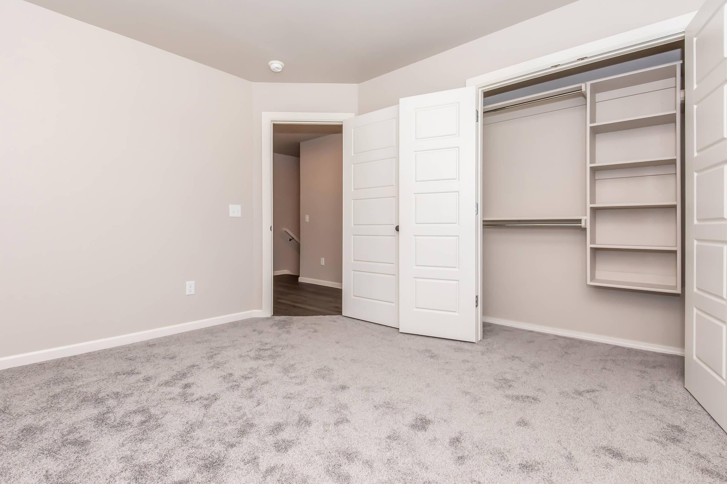 Empty room with light beige walls, soft gray carpet, and a closet with open double doors. The closet features built-in shelves. A doorway is visible in the background, leading to another room, and there is a ceiling light fixture.
