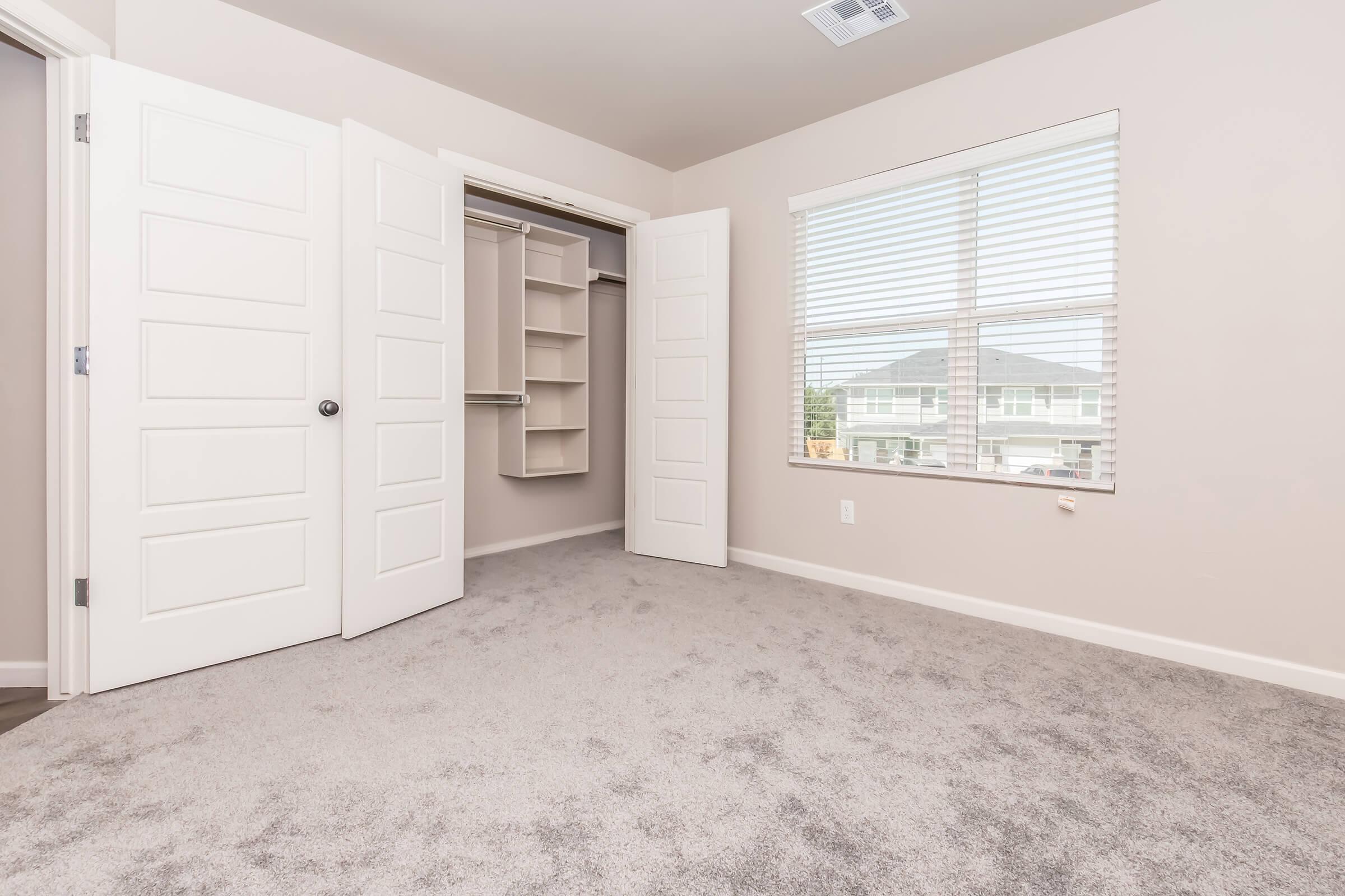 A spacious, empty room with light beige walls and soft carpet. To the left, there are two white doors leading to a closet with shelving. A large window on the right allows natural light to enter, featuring horizontal blinds and a view of the outdoors.