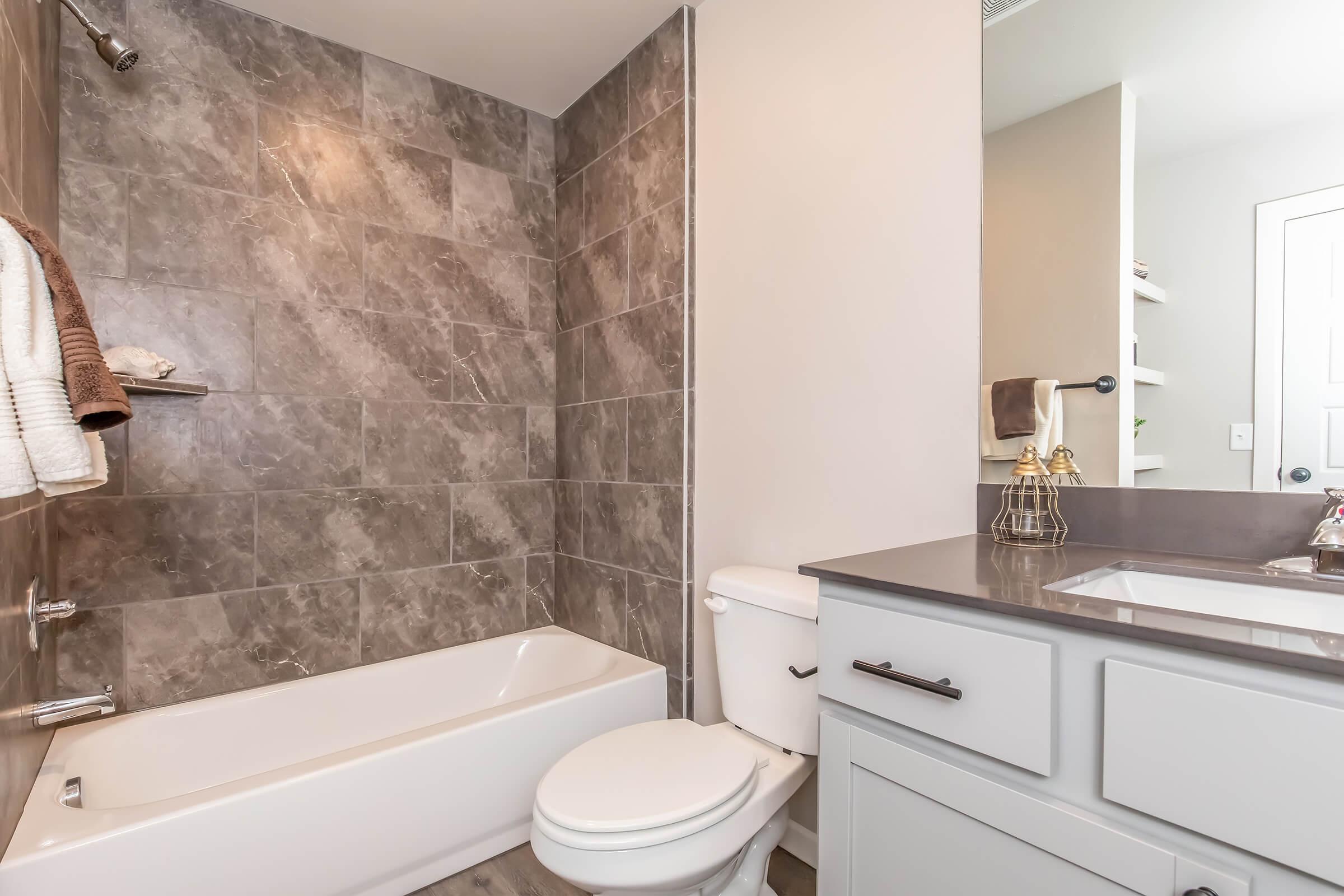 A modern bathroom featuring gray tiled walls, a white bathtub, and a toilet. The countertop has a sink with a modern faucet, and there is a small decorative item on the counter. A towel is hanging on a rack, and shelves can be seen in the background. The overall design is clean and contemporary.