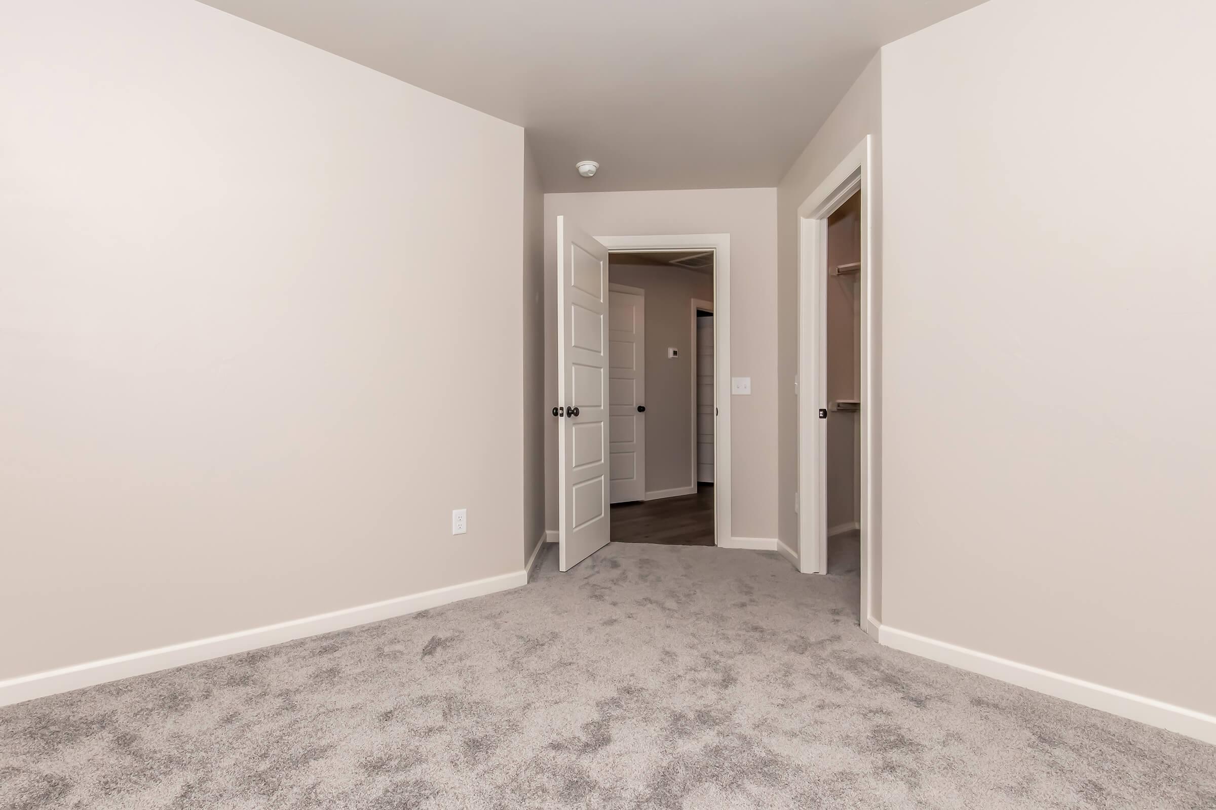A spacious, empty room with light beige walls and plush gray carpet. There are two doorways leading to adjacent areas, one of which is slightly open. The room is well-lit and has a minimalist design, creating a fresh and inviting atmosphere.