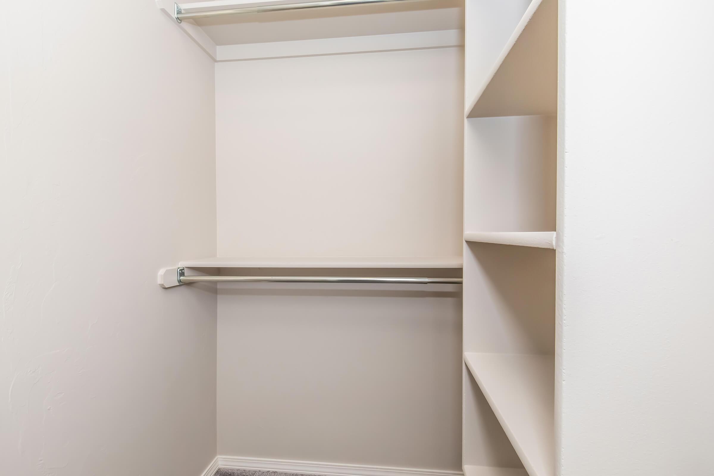 A clean, empty closet featuring white walls, a single hanging rod, and open shelving units. The floor is carpeted, and the overall space is well-lit, emphasizing the minimalist design.