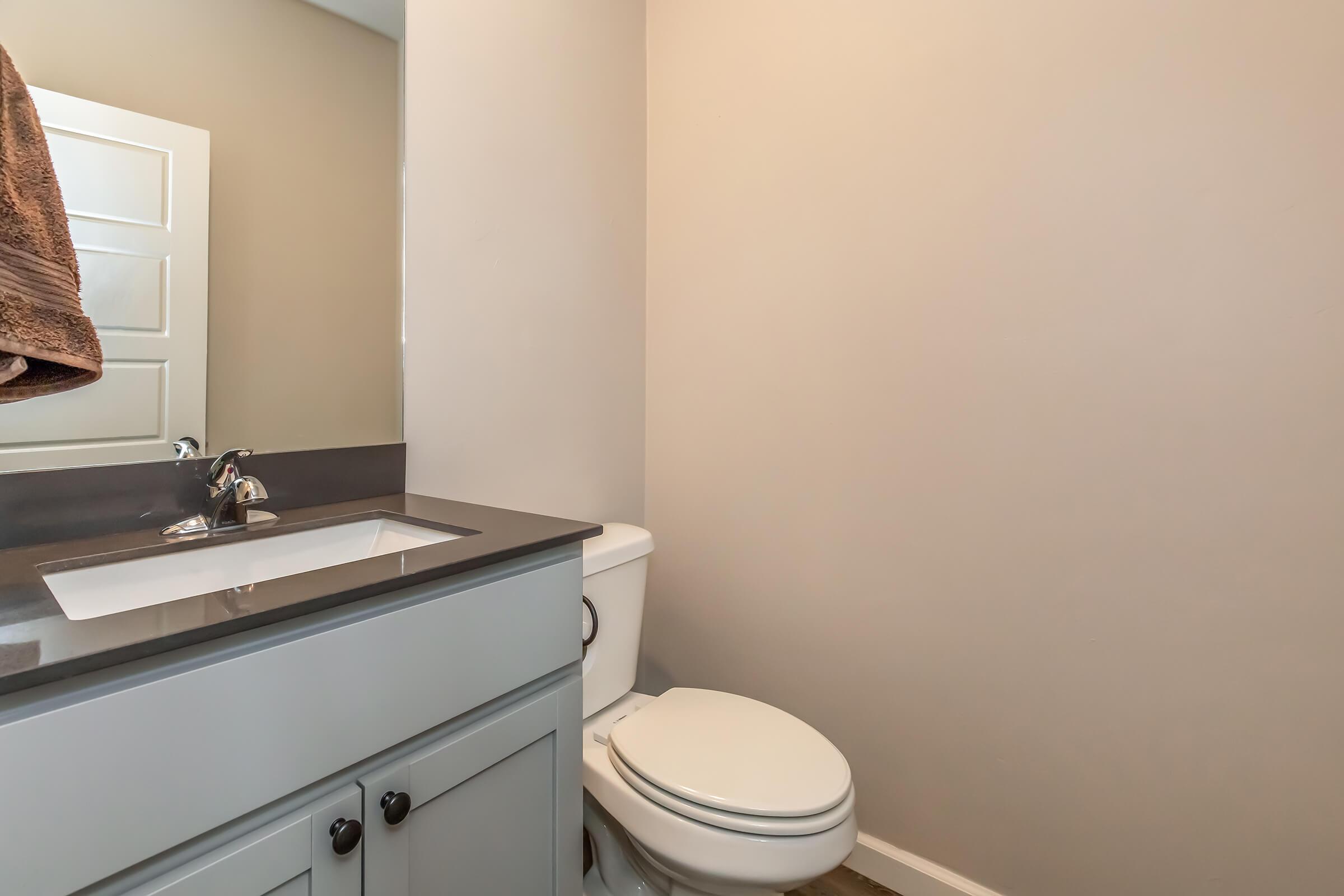 A modern bathroom featuring a toilet, a gray countertop with a sink, and a mirror. The walls are painted a light neutral color, and a brown towel hangs on a hook. The flooring is made of light-colored wood or laminate.