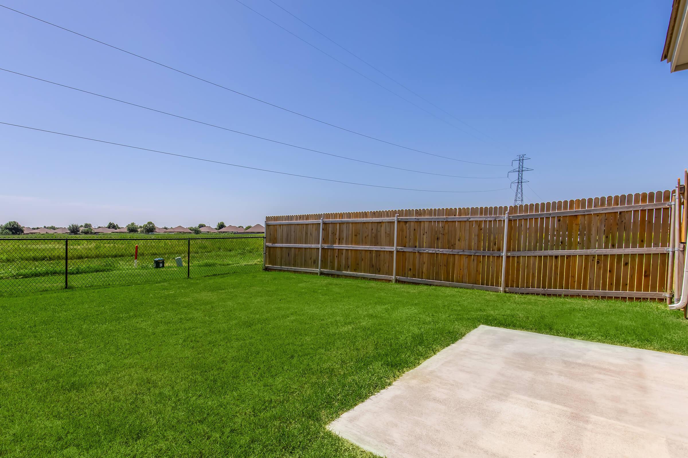 A bright, clear sky over a spacious backyard featuring lush green grass. A wooden fence runs along one side, with an adjacent chain-link fence. In the distance, power lines and a neighborhood can be seen beyond the fence. A concrete patio is visible in the foreground.