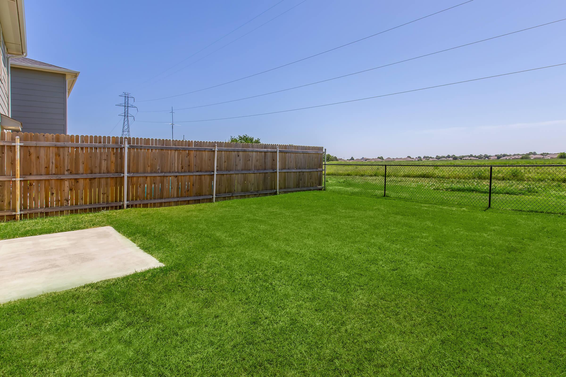 A spacious, well-maintained backyard featuring lush green grass. A concrete patio extends from the house, with a wooden fence lining one side and a chain-link fence surrounding the area. In the background, a clear blue sky meets an open field. Power lines are visible above the fence.