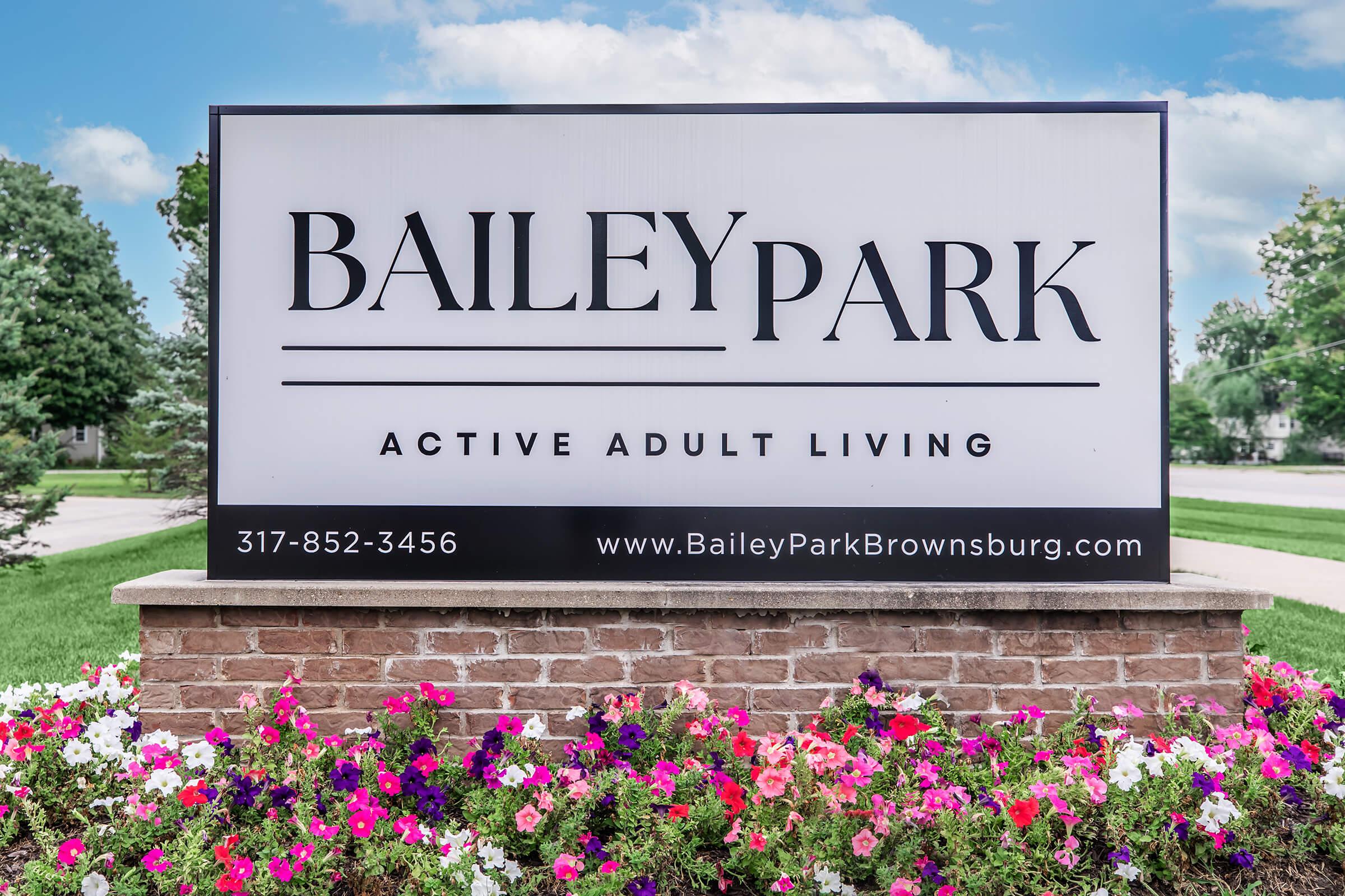 Sign for Bailey Park, a community offering active adult living. Features contact number 317-852-3456 and website www.BaileyParkBrownsburg.com. Surrounded by a landscaped area with colorful flowers in bloom.