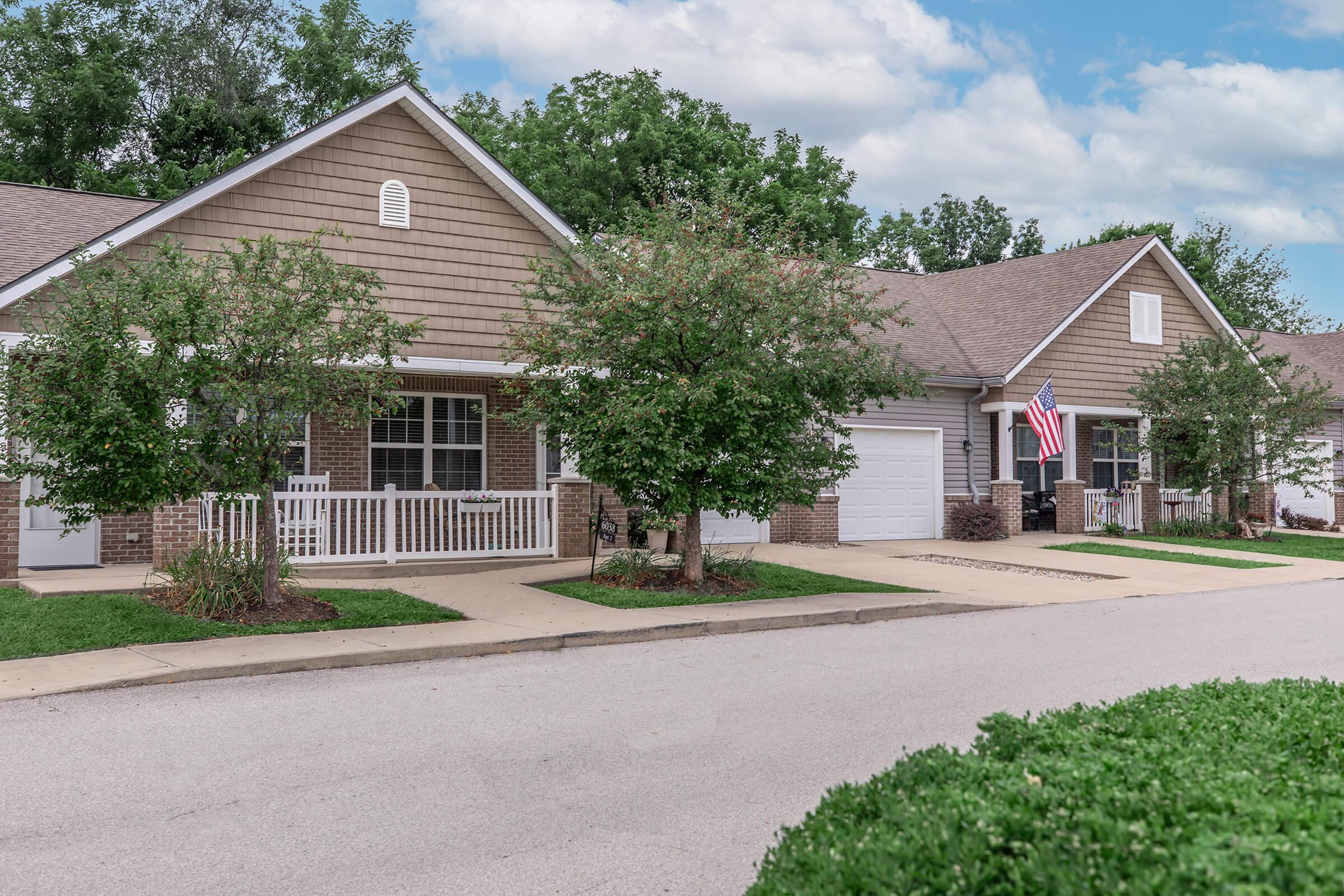 A peaceful residential street scene featuring several single-story homes with brown siding and white porches. Lush green trees and shrubs line the sidewalk, and one house displays an American flag. The sky is partly cloudy, adding to the serene suburban atmosphere.
