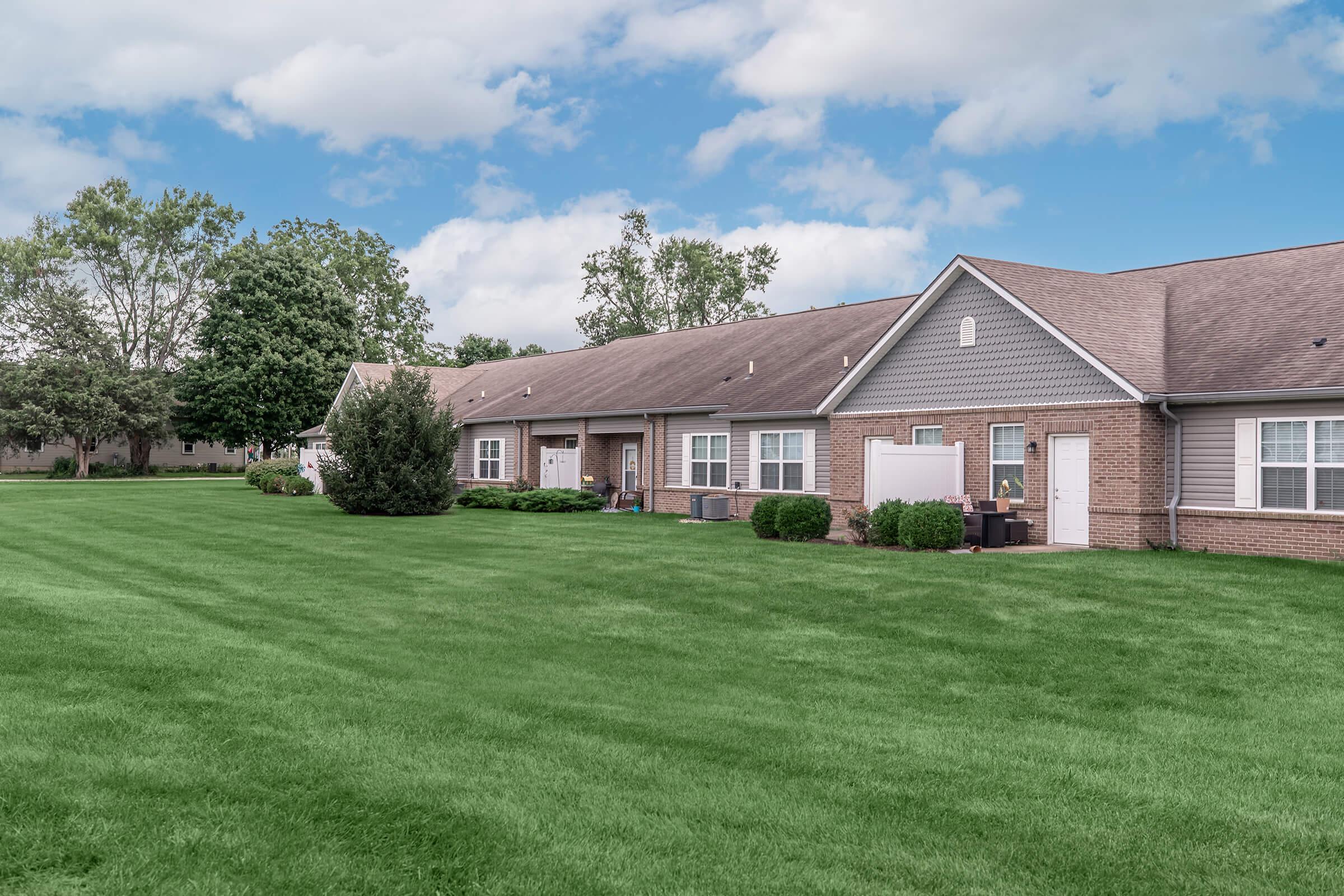 A row of brick apartment buildings with gray roofs, situated on a well-maintained lawn. The sky is partly cloudy, and there are trees in the background, creating a peaceful residential setting.