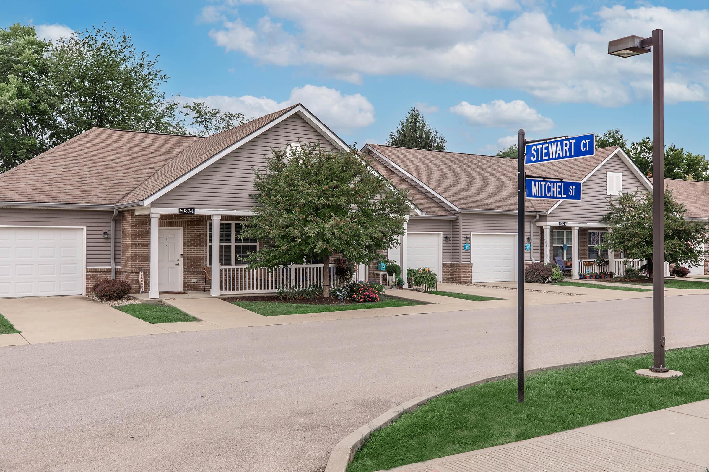 A residential street scene featuring two single-story houses with neatly manicured lawns and driveways. A blue street sign indicates the intersection of Stewart Court and Mitchel Street. The sky is partly cloudy, adding to the suburban atmosphere.