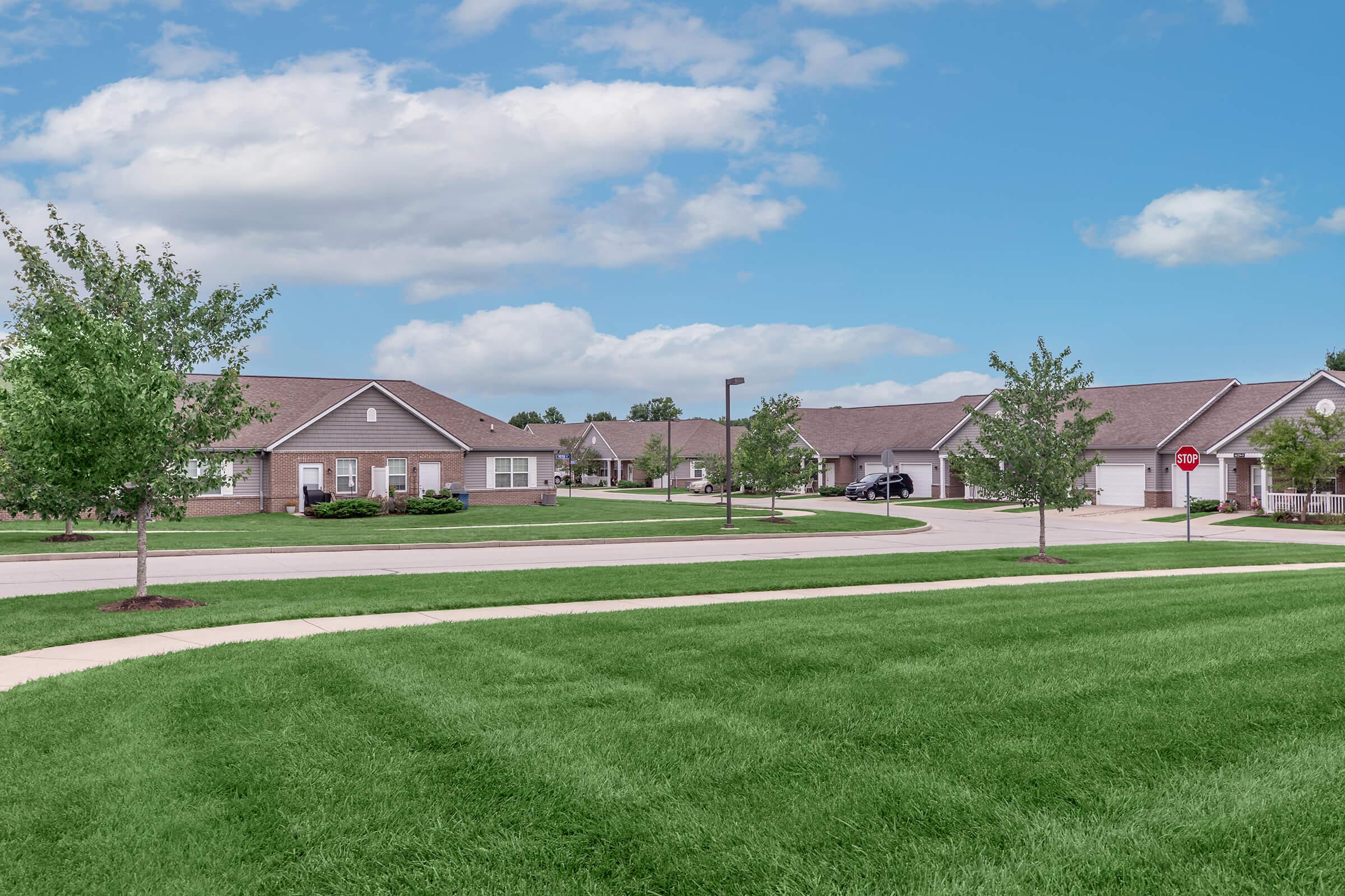 A peaceful suburban neighborhood featuring well-maintained single-story homes. Green lawns and young trees line the streets under a partly cloudy blue sky. A stop sign is visible along the curved sidewalk, adding to the community feel. The area appears calm and inviting, ideal for families or retirees.