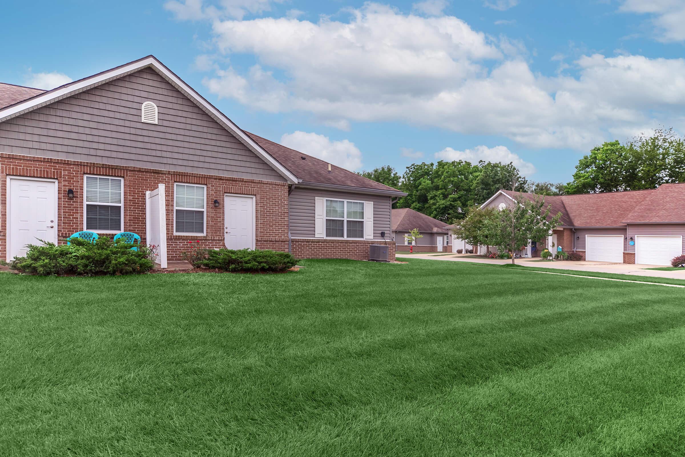 Residential neighborhood featuring a well-maintained grassy lawn with several single-story brick homes. The sky above is partly cloudy, and some homes are visible in the background, showcasing a peaceful suburban setting.