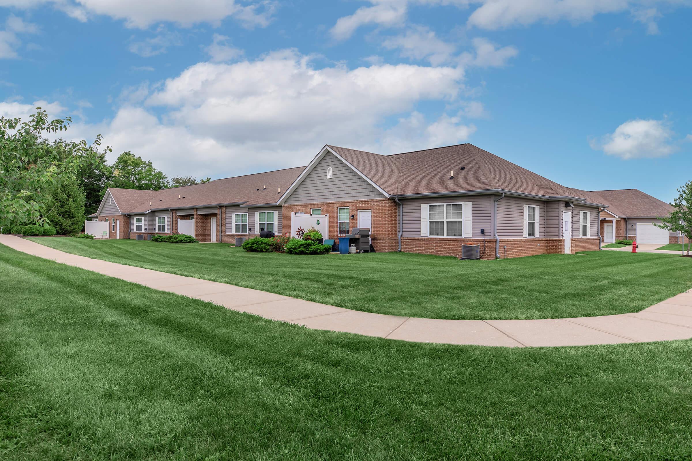 A row of single-story apartment buildings with light gray siding and brown roofs. The buildings are surrounded by green lawns and neatly trimmed bushes. A paved walkway curves through the grass, leading to the entrance of the apartments under a blue sky with fluffy white clouds.