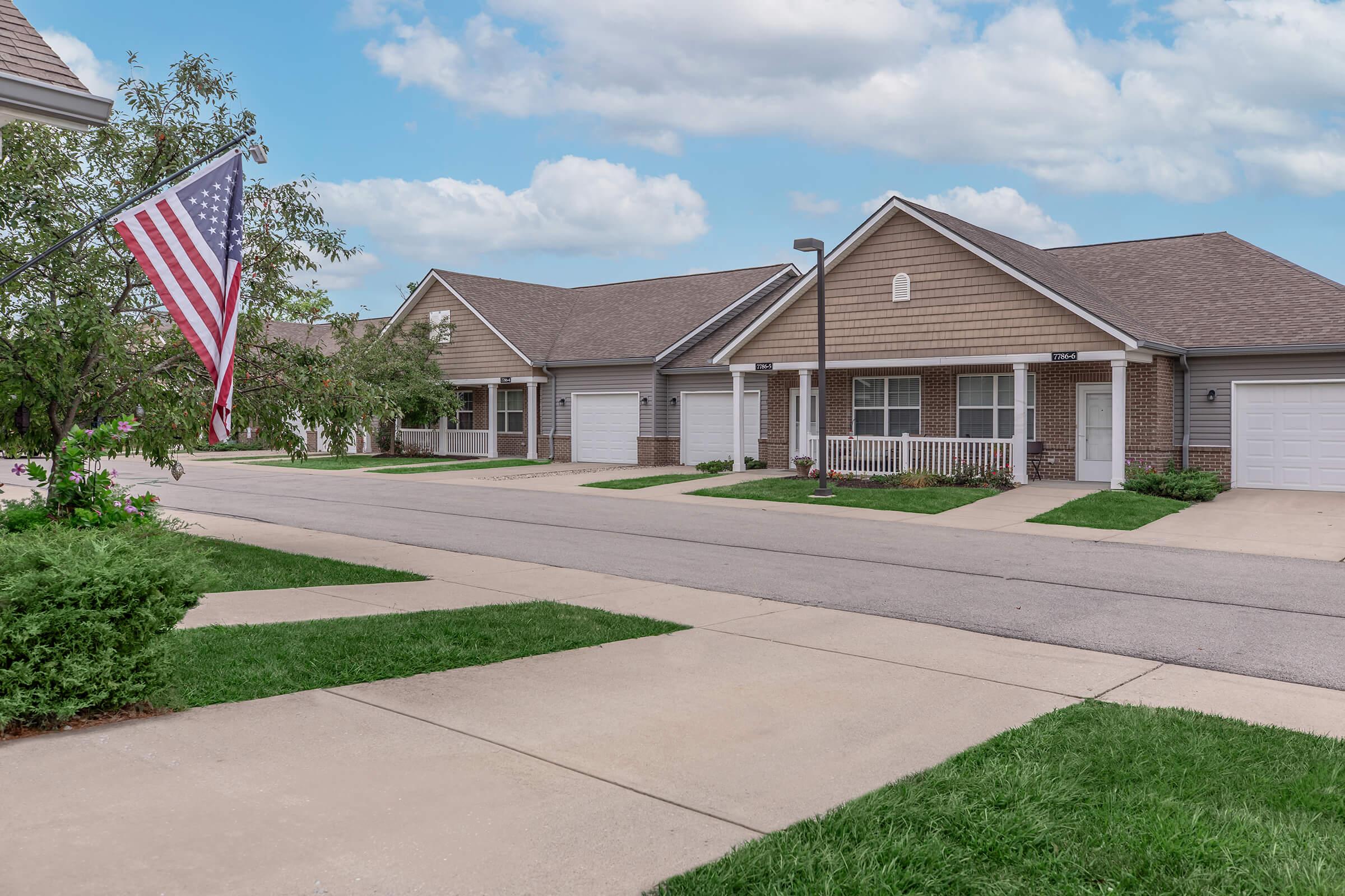 A quiet residential street featuring several single-story homes with beige siding. Each house has a front porch and a garage. An American flag is displayed on a pole in the foreground. The area is green with grassy patches and small trees, under a partly cloudy sky.