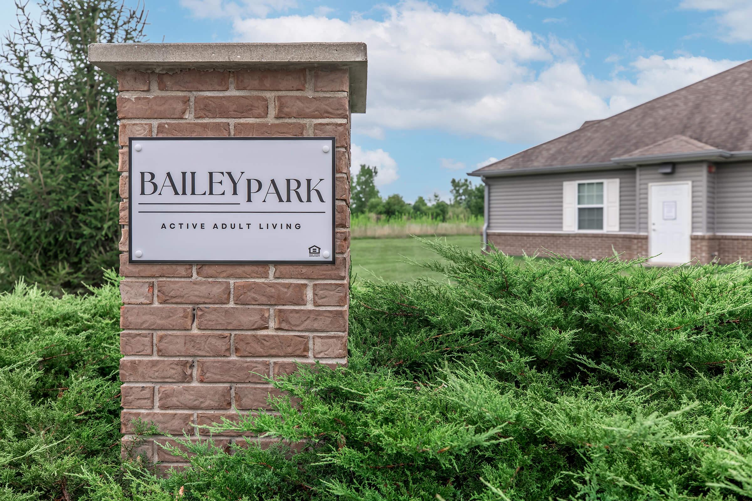 A sign marked "BAILEYPARK" indicating "ACTIVE ADULT LIVING," located on a brick pillar surrounded by greenery, with a residential building visible in the background under a partly cloudy sky.