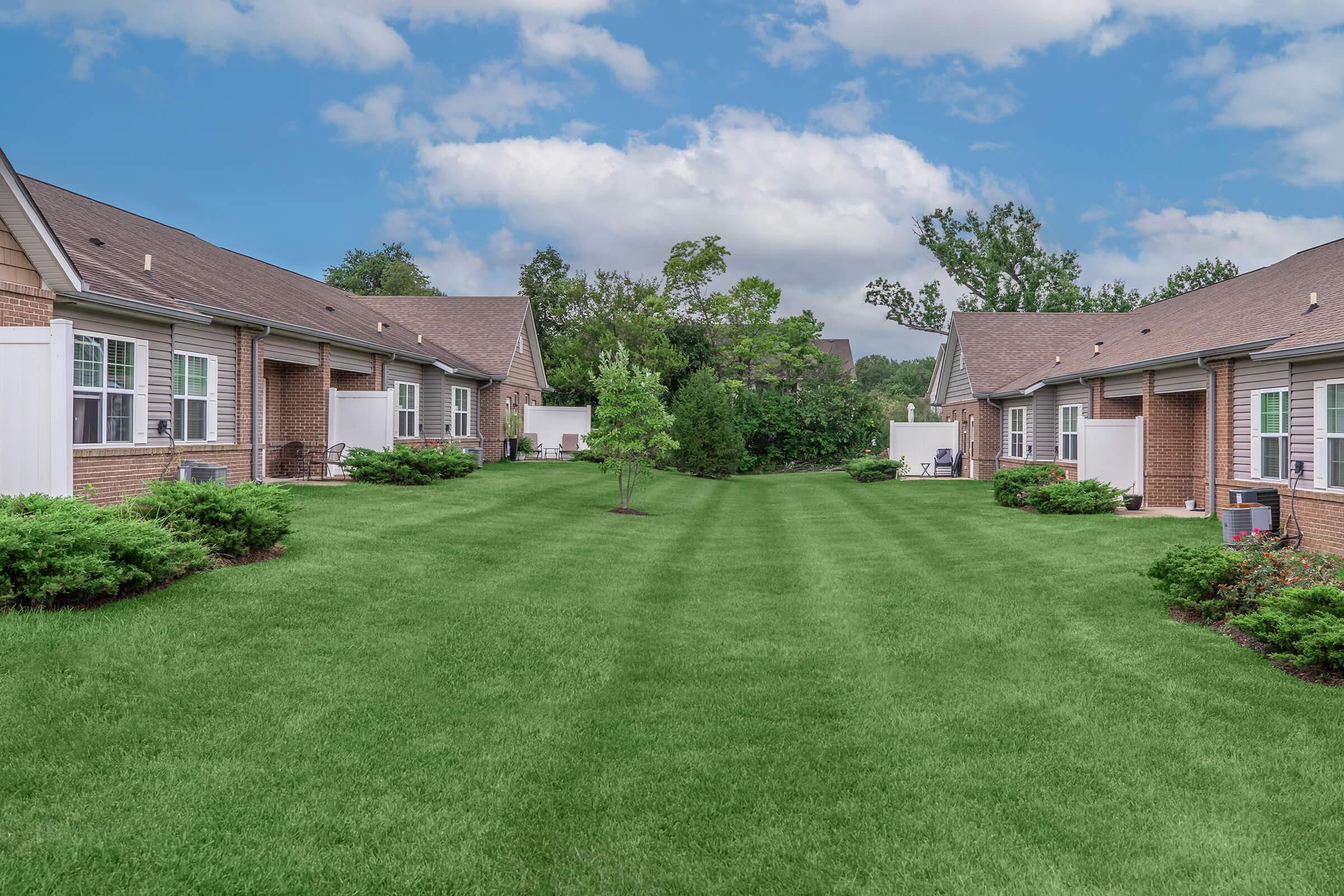 A serene view of a well-maintained residential complex featuring two sets of brick buildings. In between, a lush green lawn is divided by neatly trimmed grass, surrounded by small bushes and trees. The sky above is partly cloudy, enhancing the peaceful ambiance of the outdoor space.