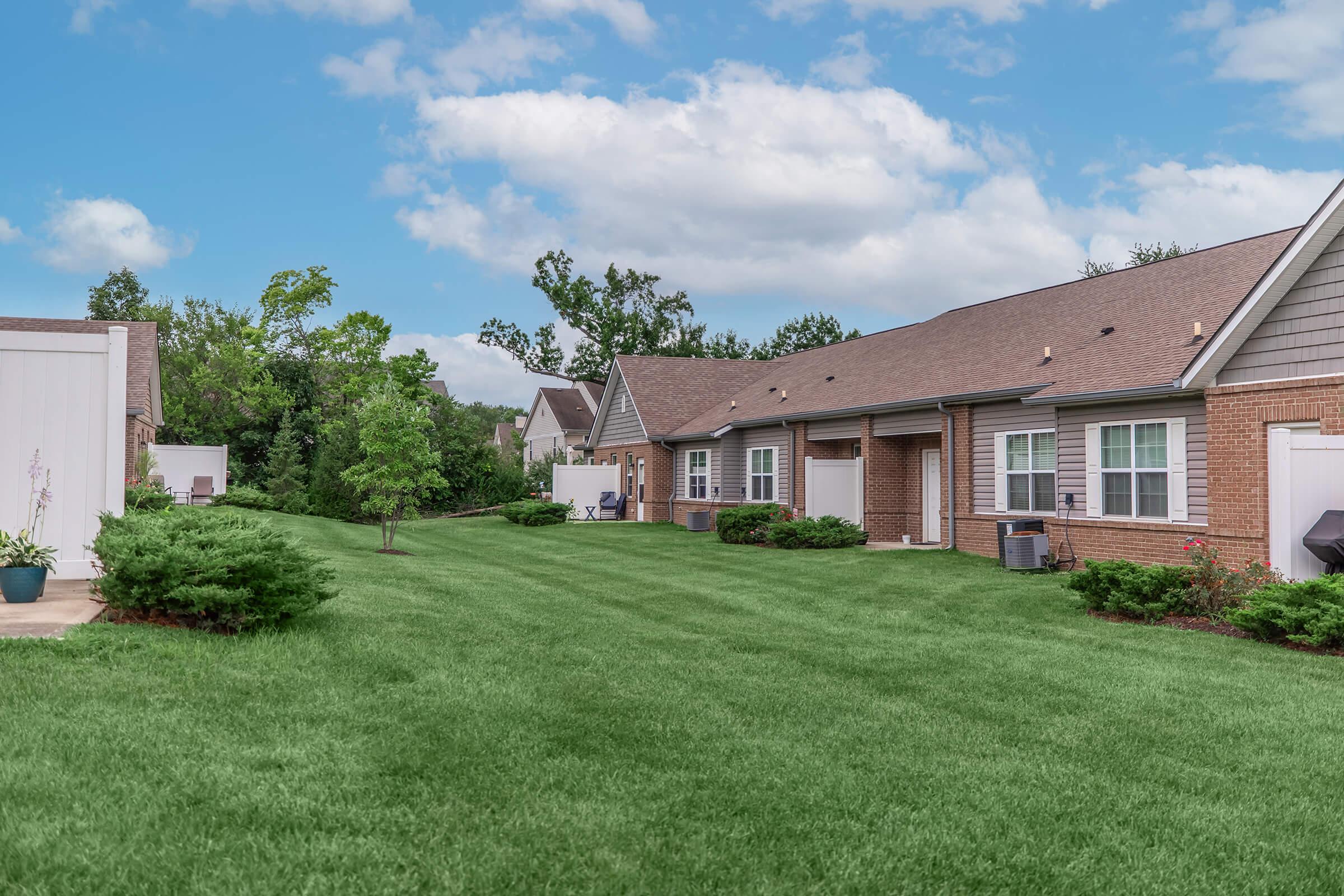 A spacious green lawn in a residential area, featuring a row of brick buildings with multiple units. The sky is partly cloudy, and trees are visible in the background. Well-maintained shrubs and small plants line the edges of the lawn, creating a neat and inviting atmosphere.