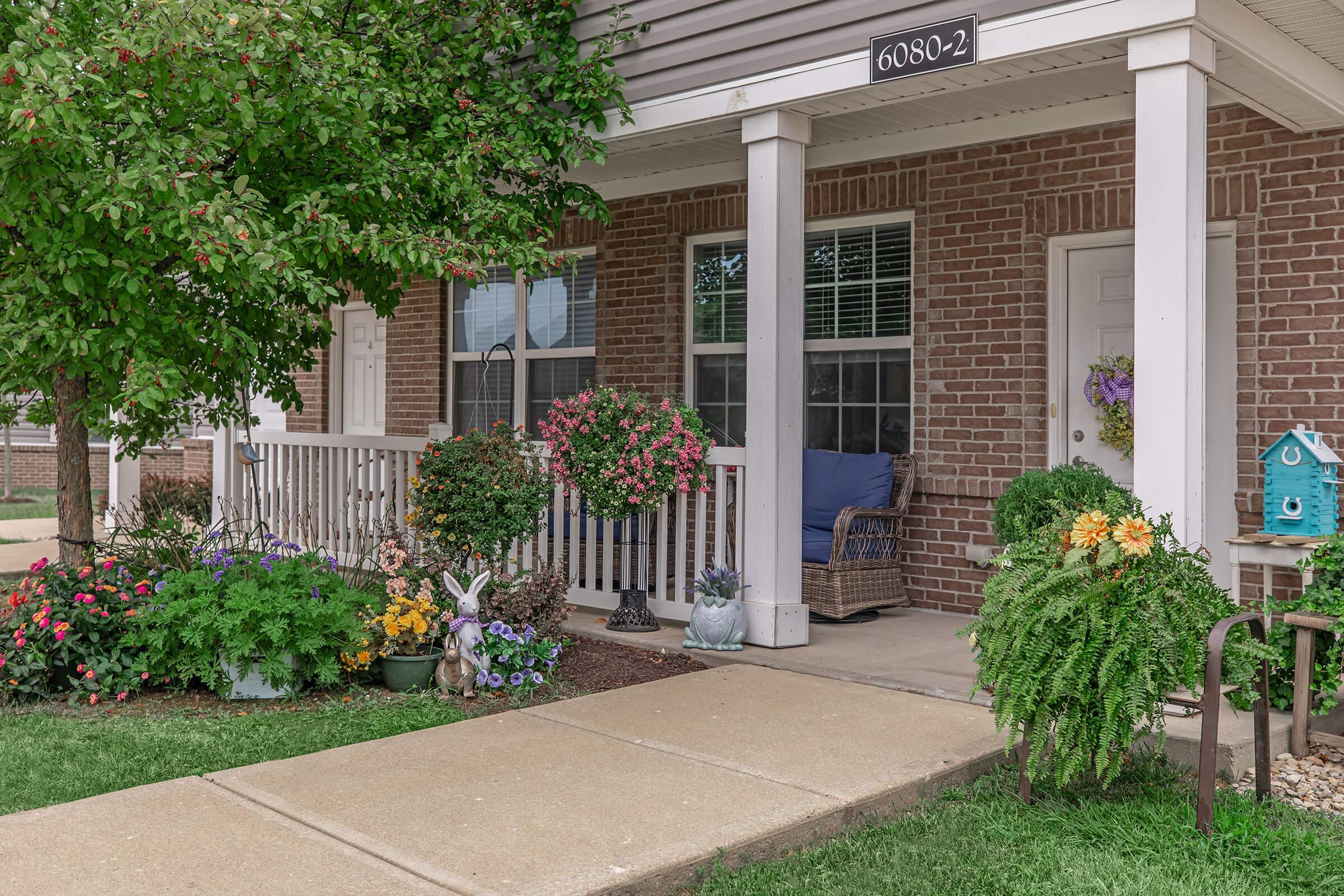 A charming front porch with a welcoming atmosphere, featuring colorful flower pots, a wicker chair, and decorative plants. The entrance has a flower-filled garden, a small blue birdhouse, and a whimsical bunny statue, creating a cheerful and inviting space for relaxation.