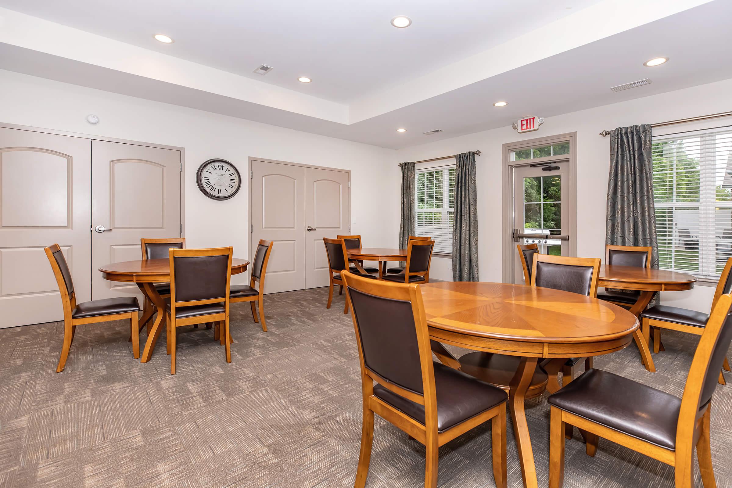 Interior view of a well-lit community room featuring several round wooden tables with brown chairs. The walls are light-colored, and there is a clock on one wall. Large windows with gray curtains allow natural light to fill the space, creating an inviting atmosphere. A door leads outside.