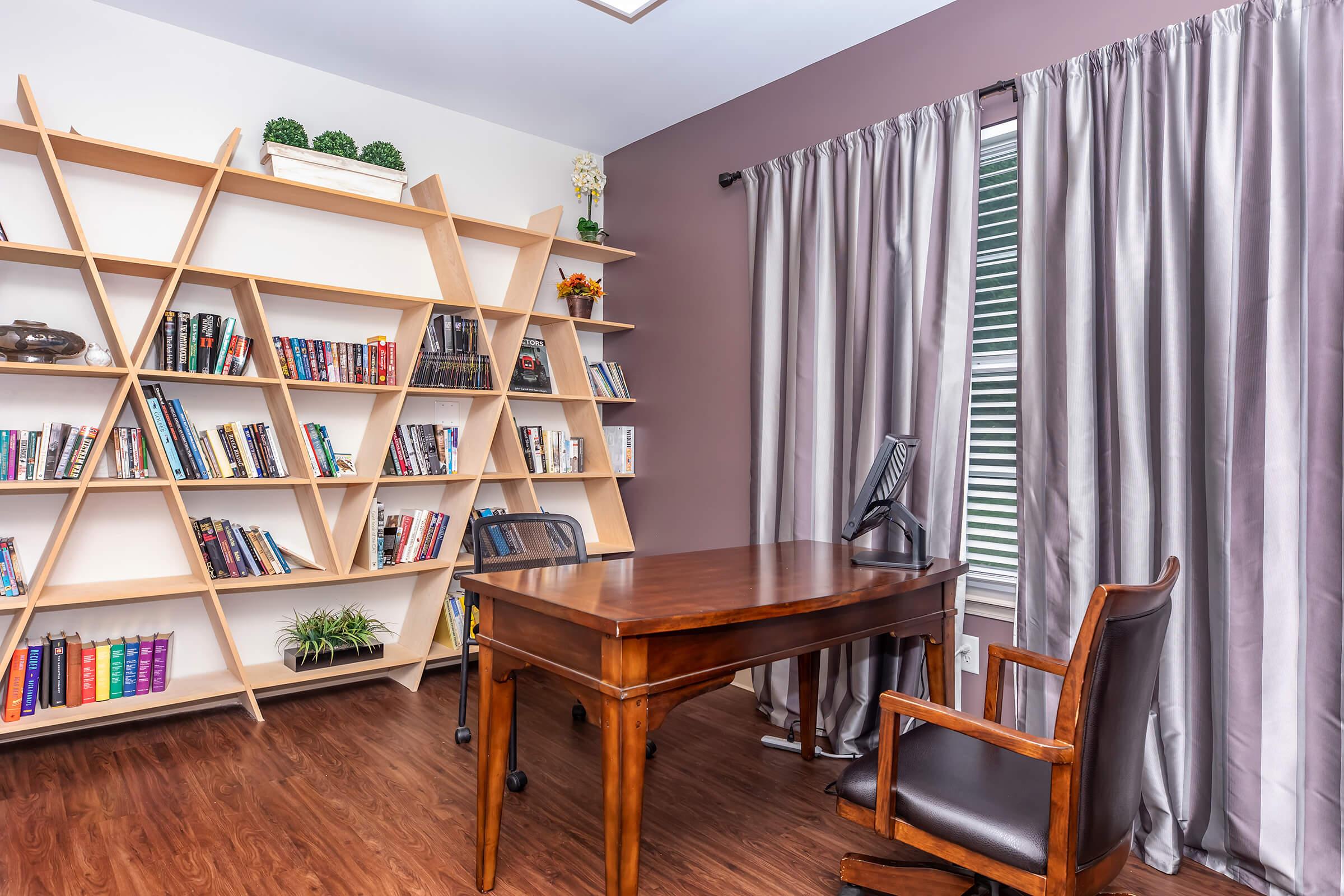 A stylish home office featuring a wooden desk and an ergonomic chair. A unique, modern bookshelf displays various books and decorative items. The walls are painted in a warm, muted tone, and there's a large window covered with elegant curtains, allowing natural light to brighten the space.