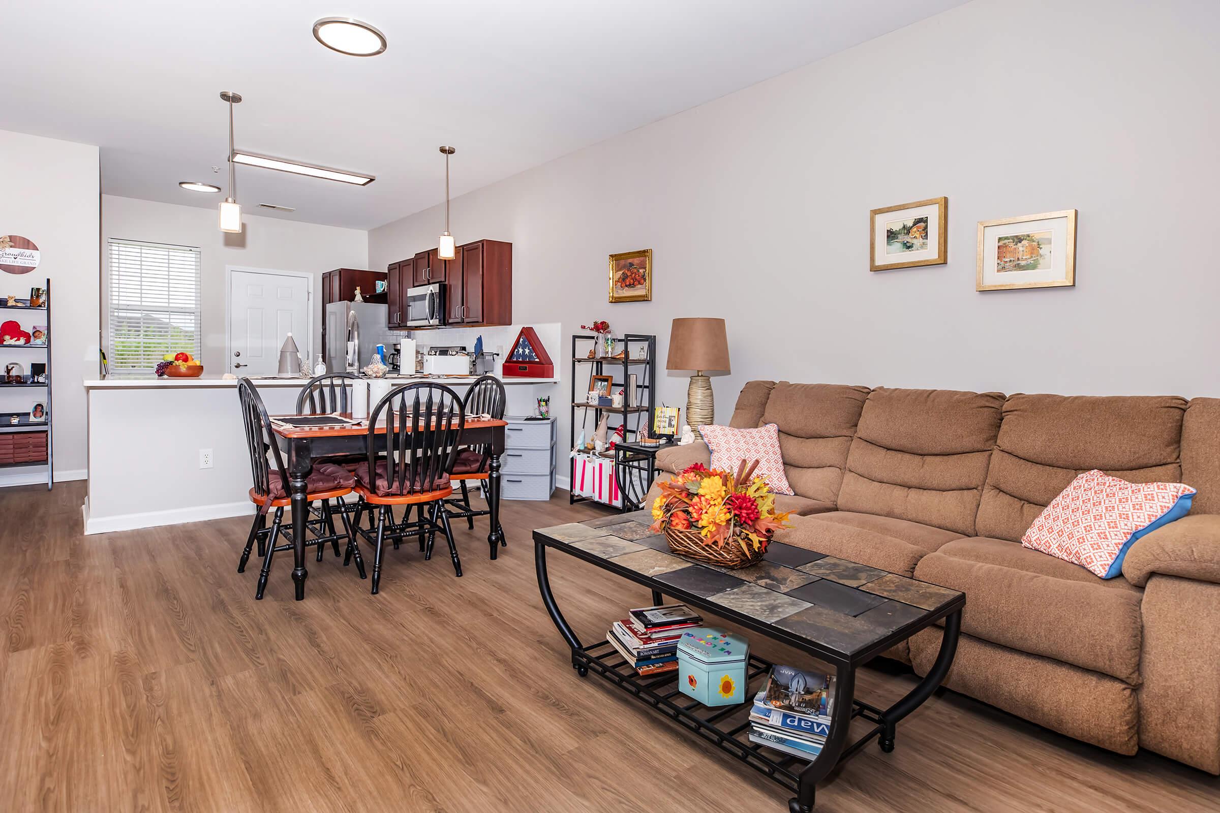 Cozy living area featuring a brown couch with decorative pillows, a wooden coffee table with books and a centerpiece, and a dining area with black chairs. The kitchen is visible in the background, with wooden cabinets and modern appliances, illuminated by pendant lights. Warm, inviting atmosphere with autumn decor.