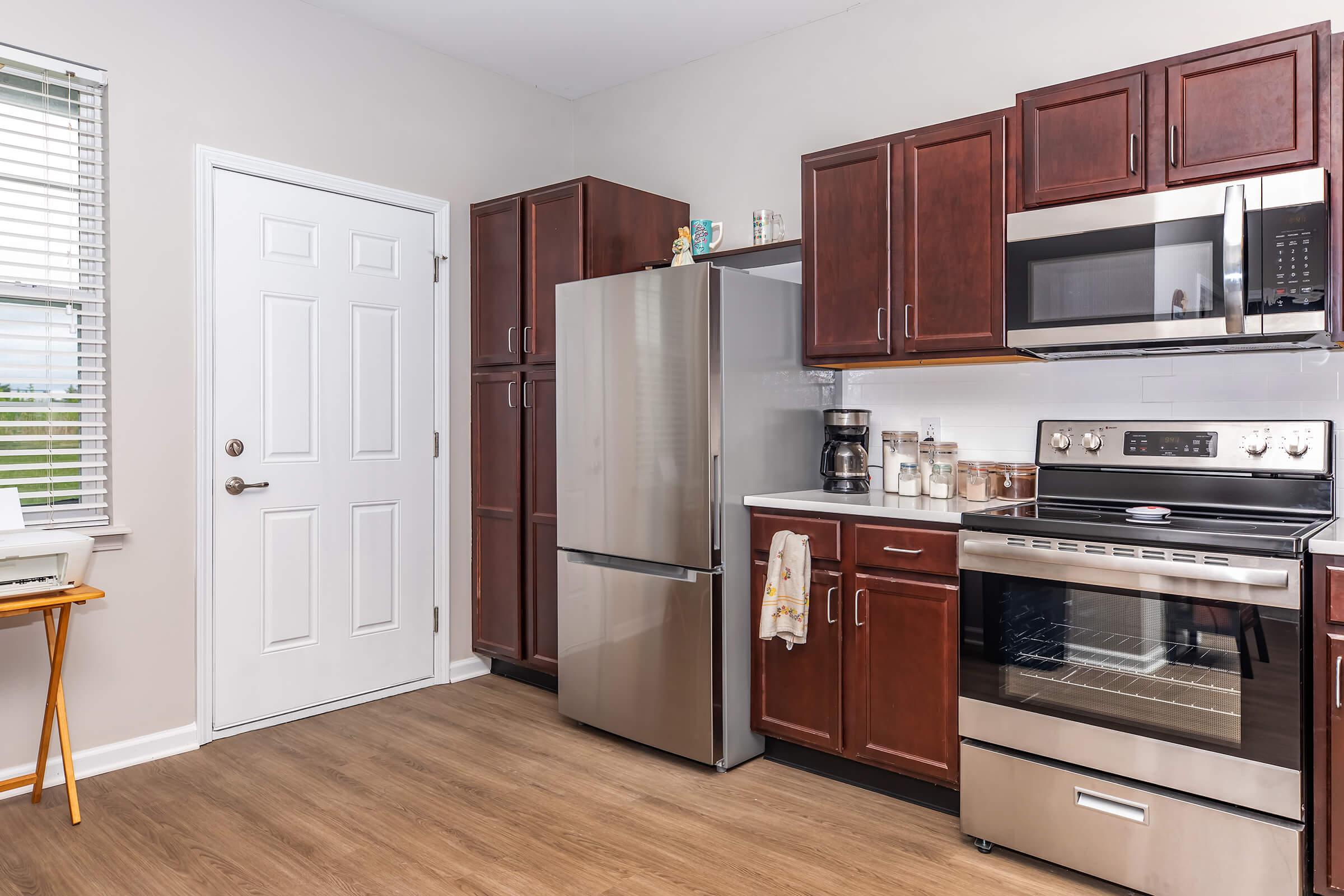 A modern kitchen featuring dark wooden cabinets, a stainless steel refrigerator, a microwave, and an oven. The room has a window with blinds, a small table, and various kitchen appliances. The walls are painted a light color, creating a bright and inviting atmosphere.