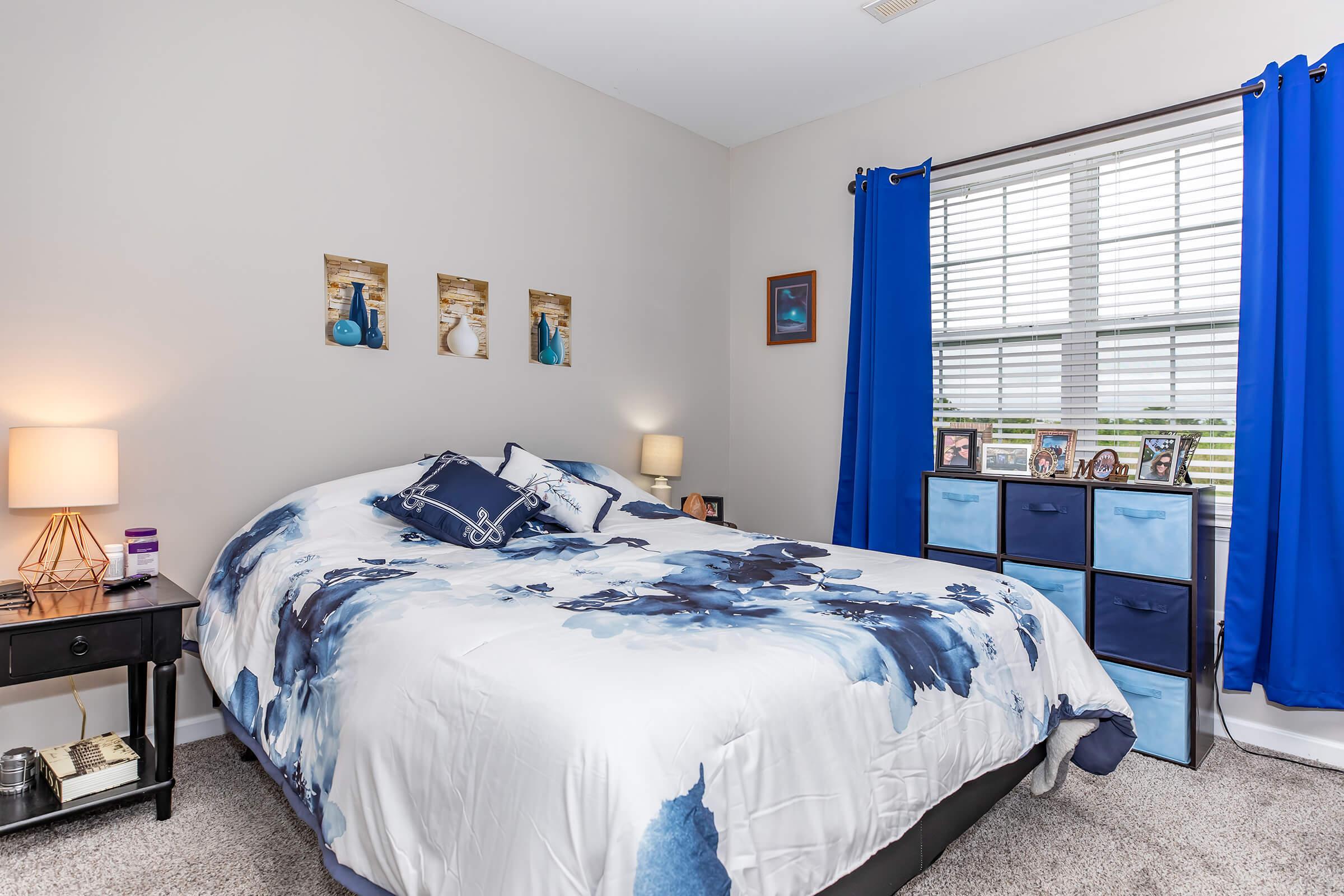 A cozy bedroom featuring a queen-sized bed with a blue and white abstract patterned comforter. There are two bedside lamps on nightstands, blue curtains at the window, and a dresser with decorative photos on top. The walls are painted in a light color, creating a welcoming and bright atmosphere.