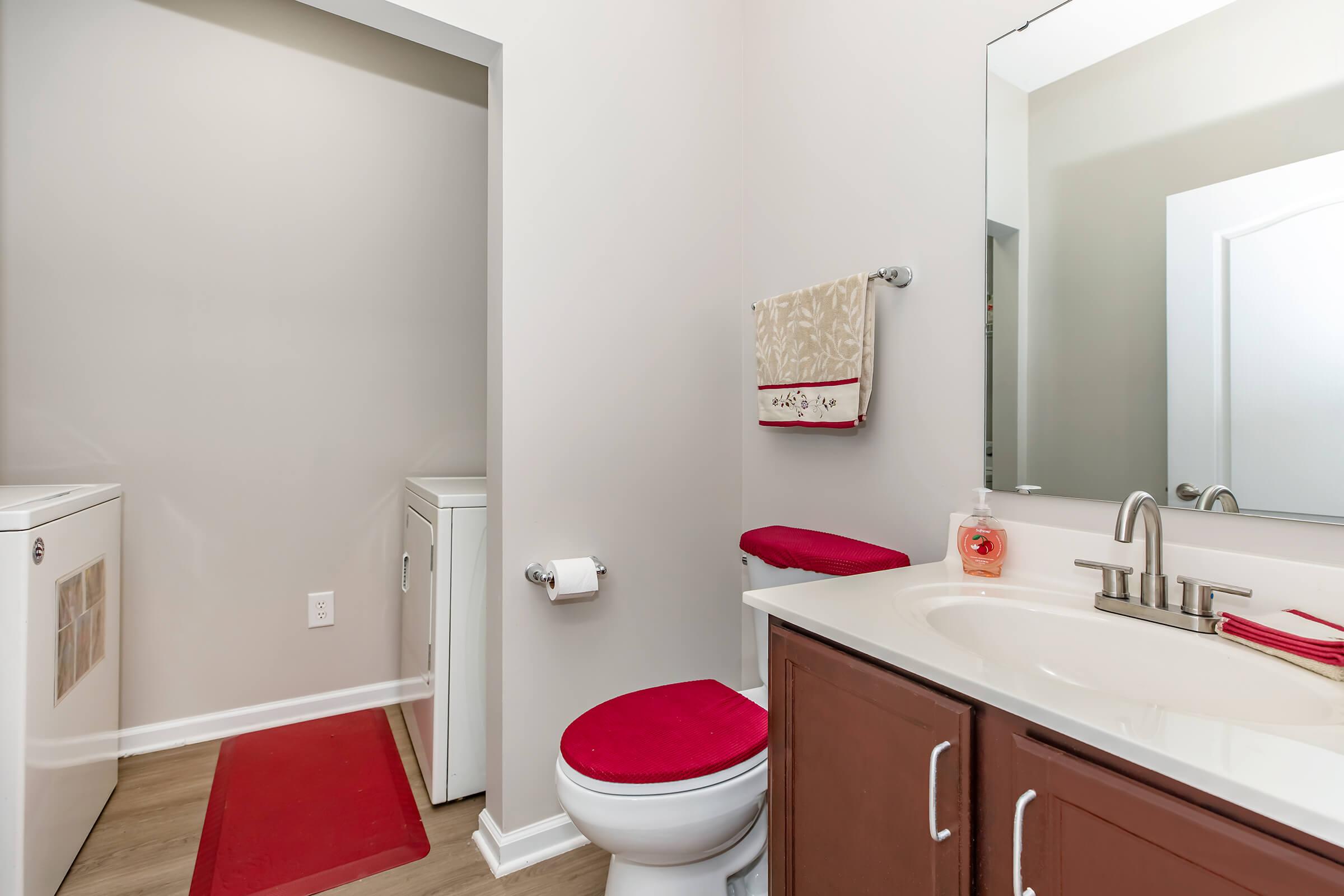 A modern bathroom featuring a white toilet, a vanity with a sink and brown cabinets, a large mirror, and red accents including a rug and towel. A laundry area is visible to the side, with a washing machine and storage space. The walls are painted in a light color, creating a bright and clean atmosphere.
