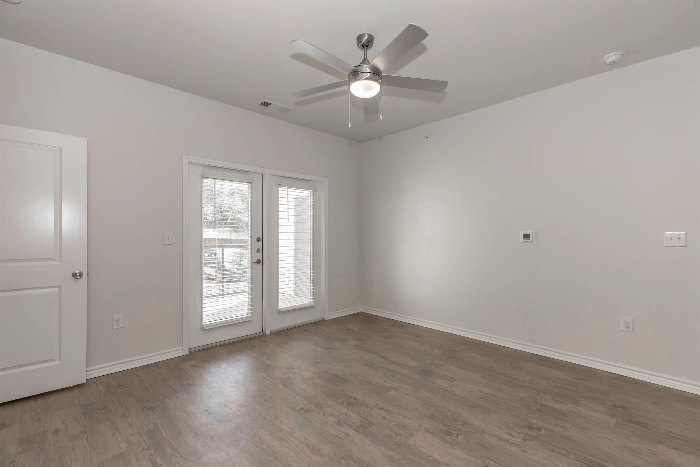 A spacious, empty room with light gray walls and wood-like flooring. It features a ceiling fan, a pair of glass doors leading to a small balcony with blinds, and a wall-mounted thermostat. The room is well-lit and has a modern, minimalist design.