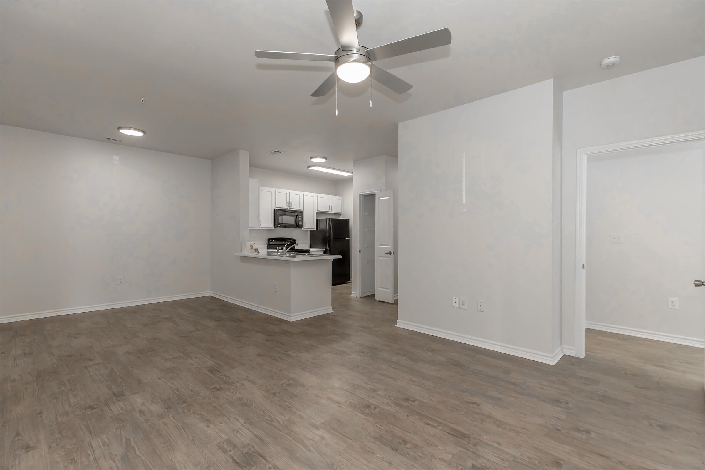 A spacious, modern living area featuring wood-style flooring, a ceiling fan, and an open layout that leads to a kitchen with white cabinets. The kitchen is partially visible, showing appliances and counter space. Two doorways suggest access to additional rooms or a bathroom. The walls are painted light colors, enhancing the bright atmosphere.