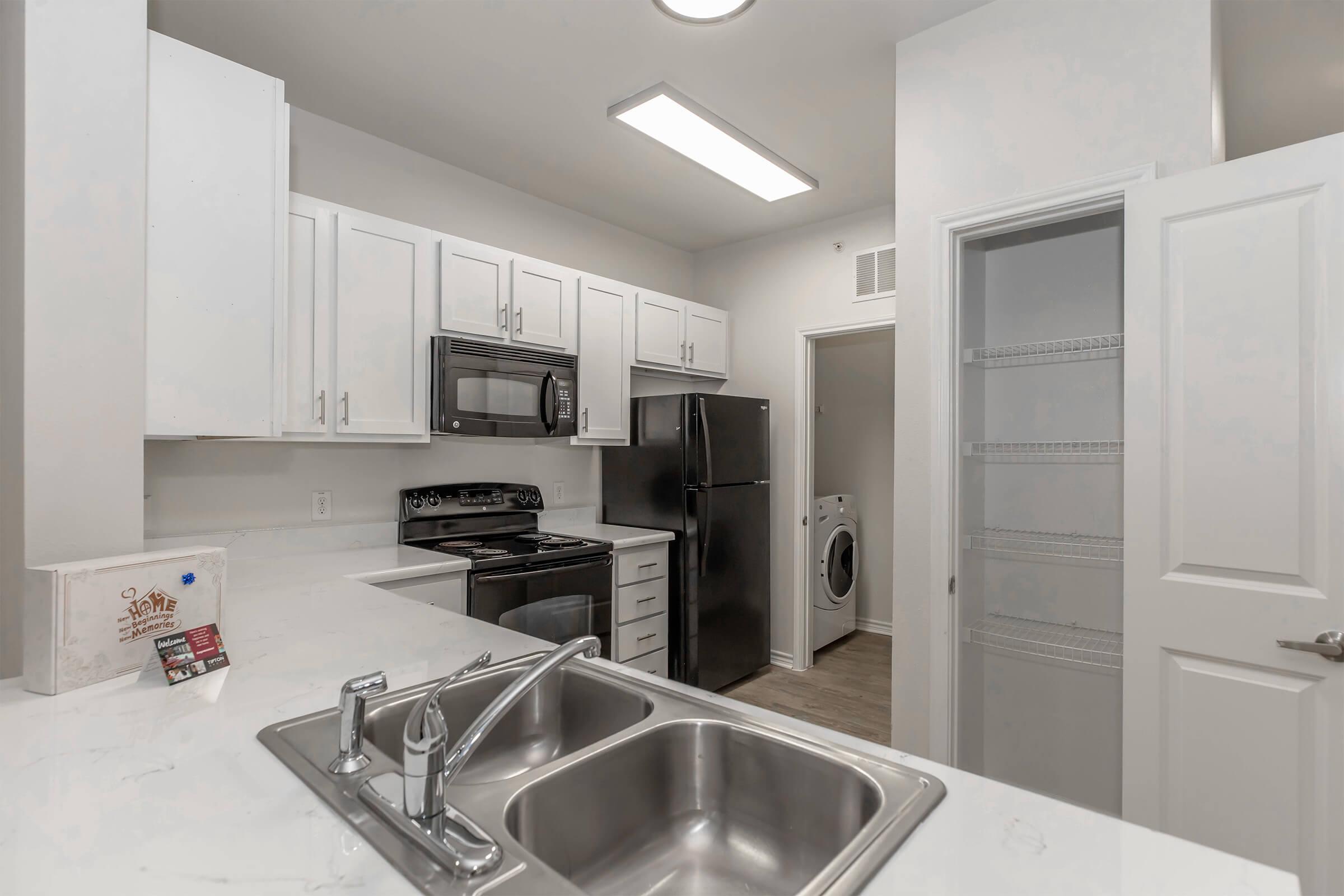 Modern kitchen featuring white cabinetry, stainless steel appliances including a microwave, stove, and refrigerator, along with a double sink. A washer and dryer are visible in a nearby closet, and the space boasts neutral-colored walls and wood-look flooring, creating a clean and functional atmosphere.
