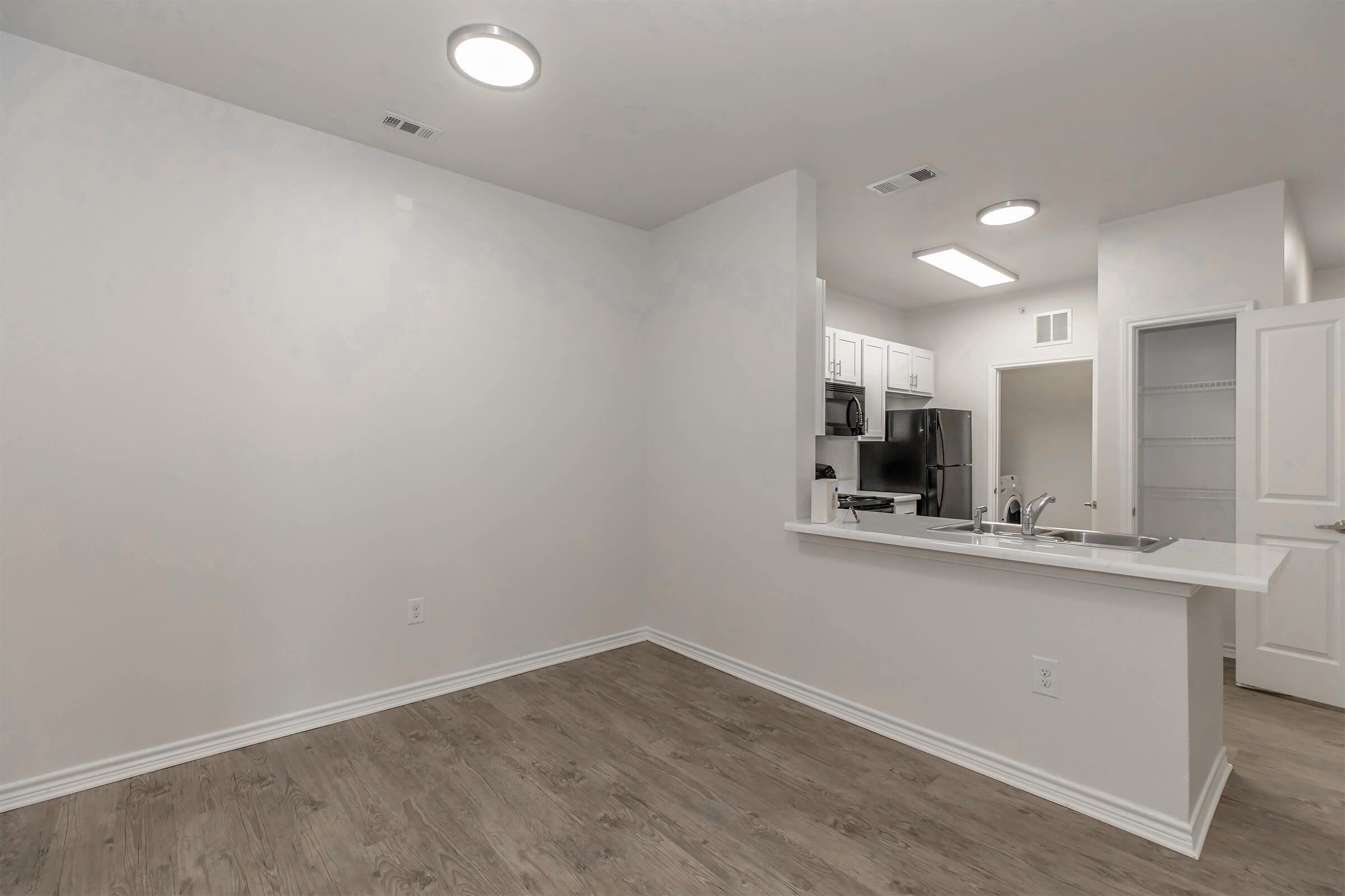 An empty, modern apartment interior featuring light-colored walls and wood-like flooring. A kitchen area with white cabinets, a black refrigerator, and a countertop is visible. Recessed lighting illuminates the space, and a door leads to another room or area.