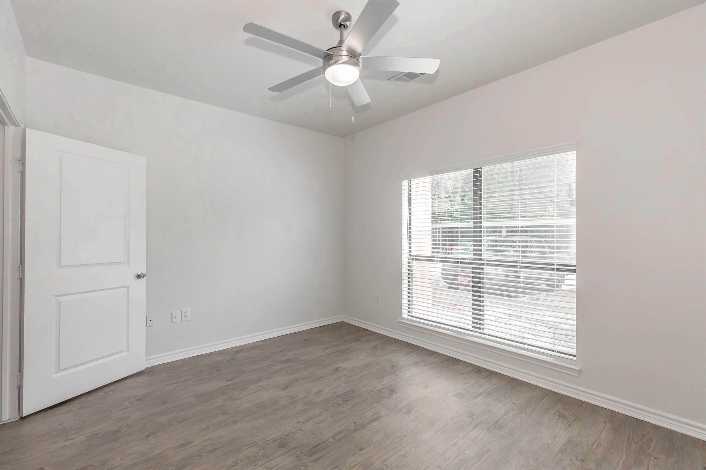 Empty room with light-colored walls and a wooden floor. A ceiling fan with five blades is mounted on the ceiling. There is a large window with blinds allowing natural light to enter, and a white door is visible on the left. The room has a clean, modern aesthetic.