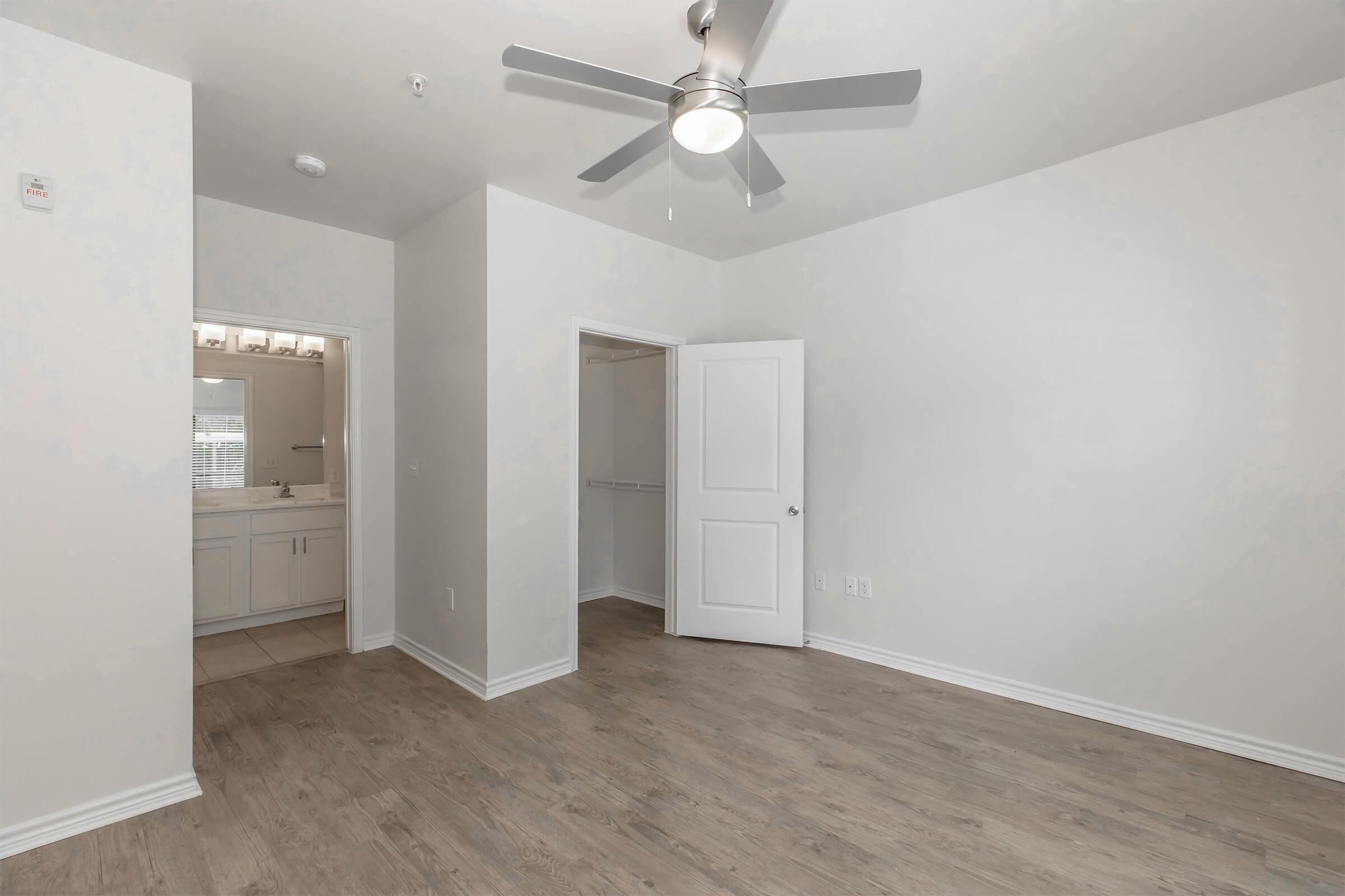 A light-colored room with a ceiling fan, wooden flooring, and a closet. A partially visible bathroom can be seen through an open doorway on the left. The walls are painted white, creating a bright and spacious atmosphere.
