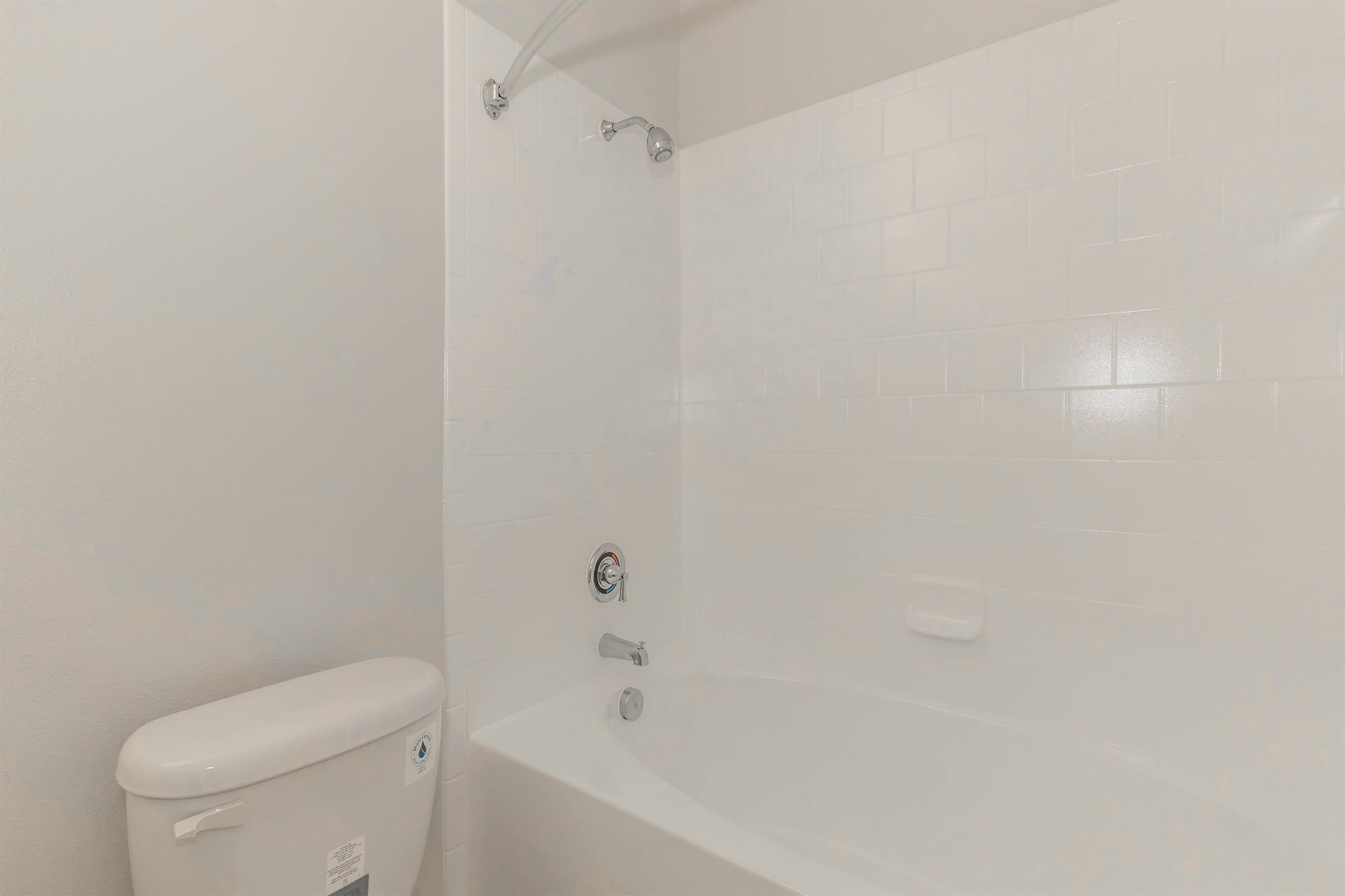 View of a clean, modern bathroom featuring a white tiled bathtub with a showerhead, a chrome faucet, and a bathtub spout. To the left, there is a white toilet. The walls are painted a light color, creating a bright and spacious atmosphere.