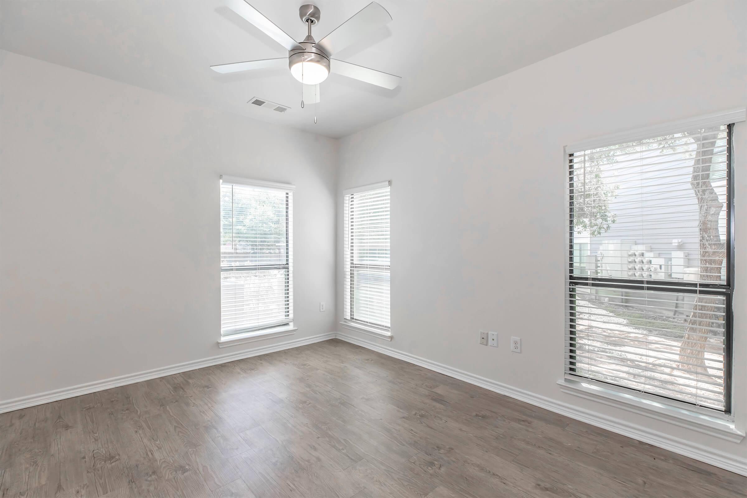 A well-lit, empty room with light-colored walls and a ceiling fan. There are two windows with blinds providing natural light, and a light wood floor. Outside the windows, greenery is visible, enhancing the room's brightness and openness. Ideal for personalizing or decorating.