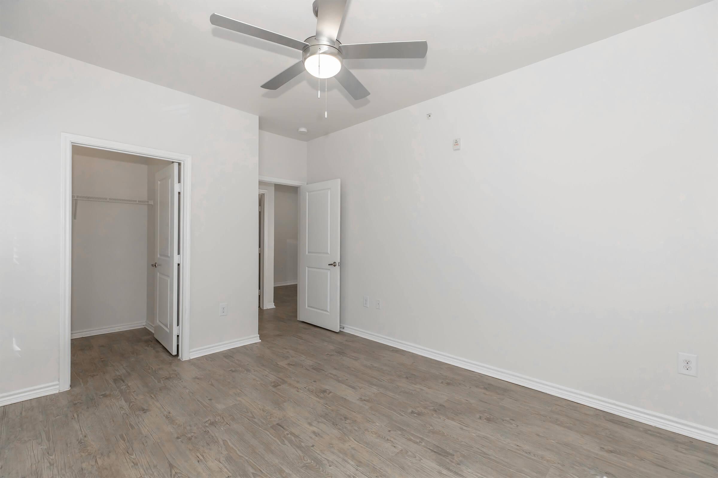 A spacious, empty room featuring light-colored walls and a ceiling fan. Two doorways lead to other areas, with one doorway opening to a small closet. The floor has a wood-like finish, contributing to a clean and modern aesthetic. Natural light fills the space, creating an inviting atmosphere.