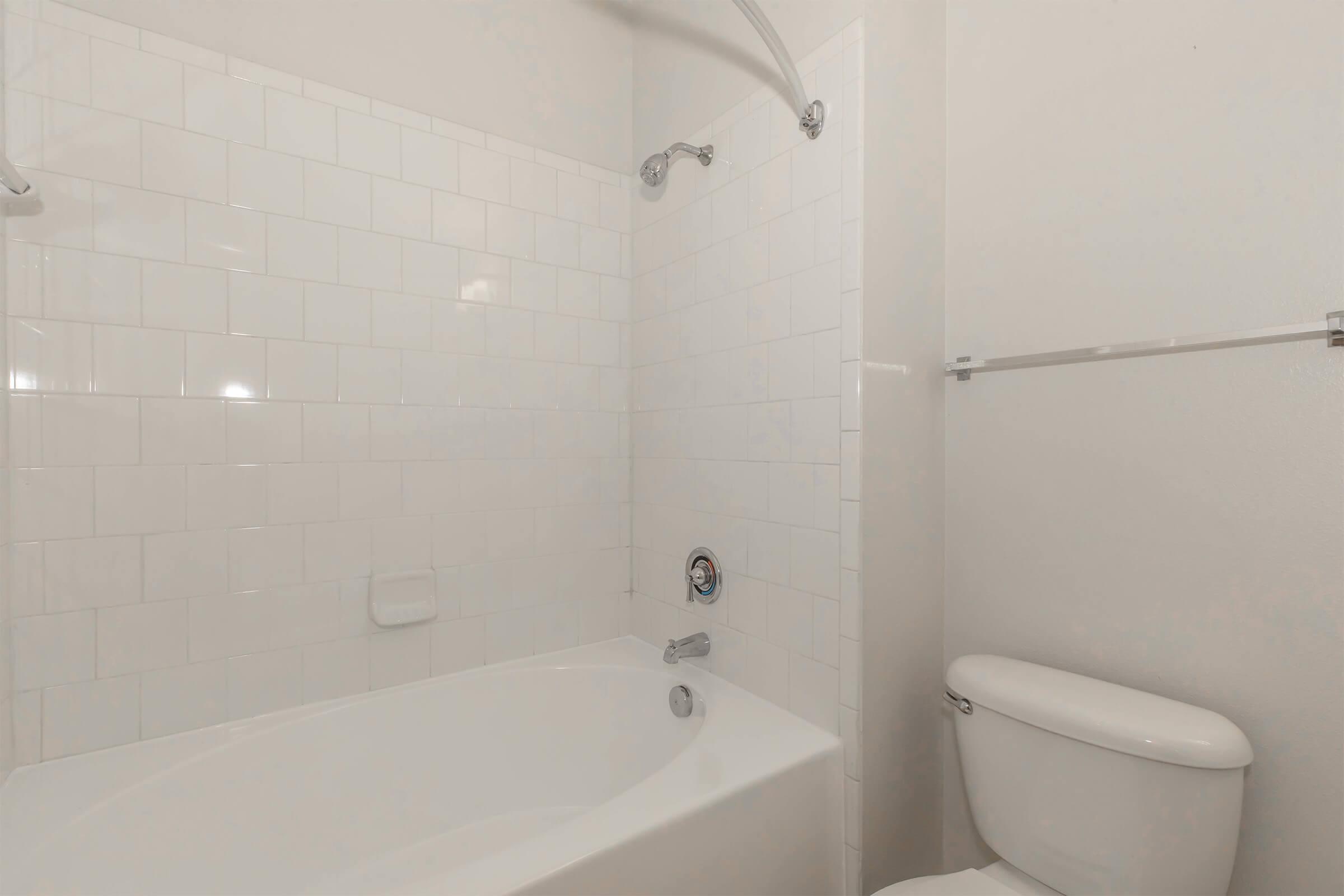 A clean bathroom featuring a white tiled shower area with a bathtub, a chrome faucet, and a wall-mounted showerhead. To the right, there is a white toilet, and the walls are painted in a light color, creating a bright and airy atmosphere.